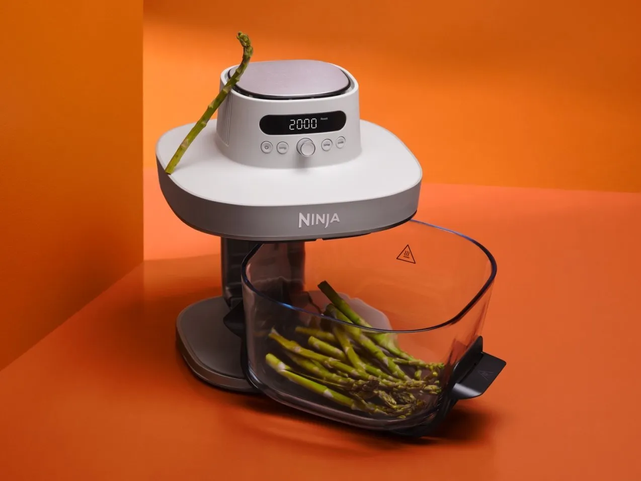 Ninja countertop steamer with digital timer and clear water tank, cooking fresh asparagus on orange background