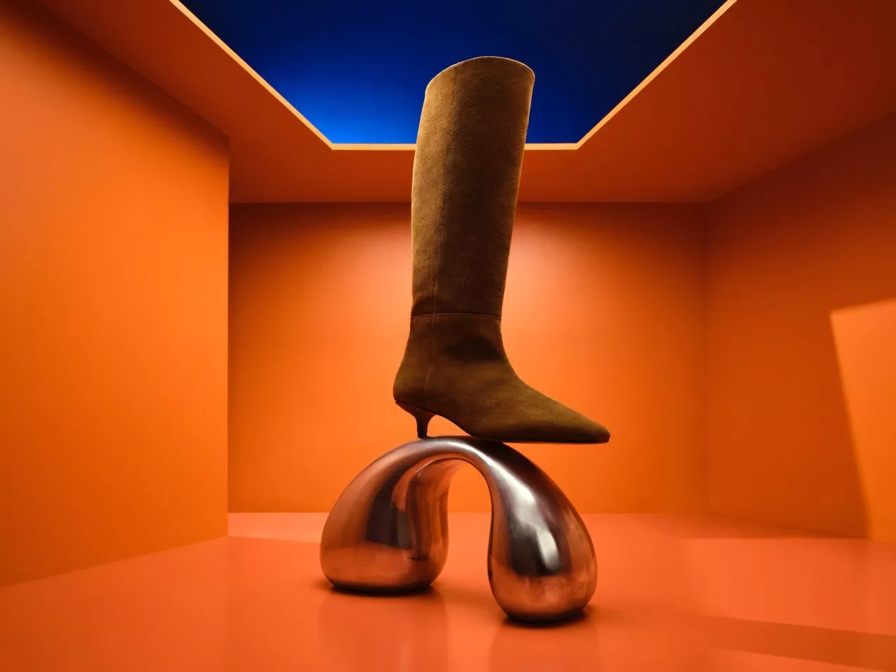 Brown suede knee-high boot on chrome sculptural stand in orange room with blue ceiling, fashion still life photo