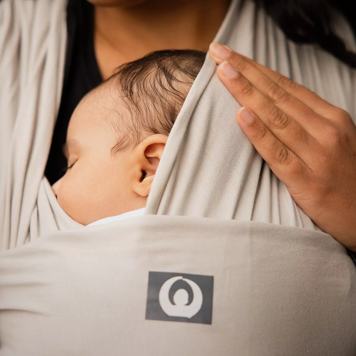 A peaceful baby sleeps in a soft grey baby wrap carrier, held close by a caring hand, showcasing comfort and security.