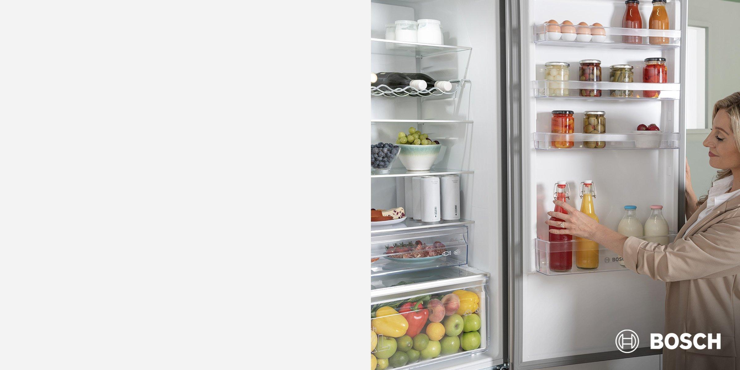 A woman organizing beverages in a well-stocked Bosch refrigerator filled with fresh produce, dairy, and jars.