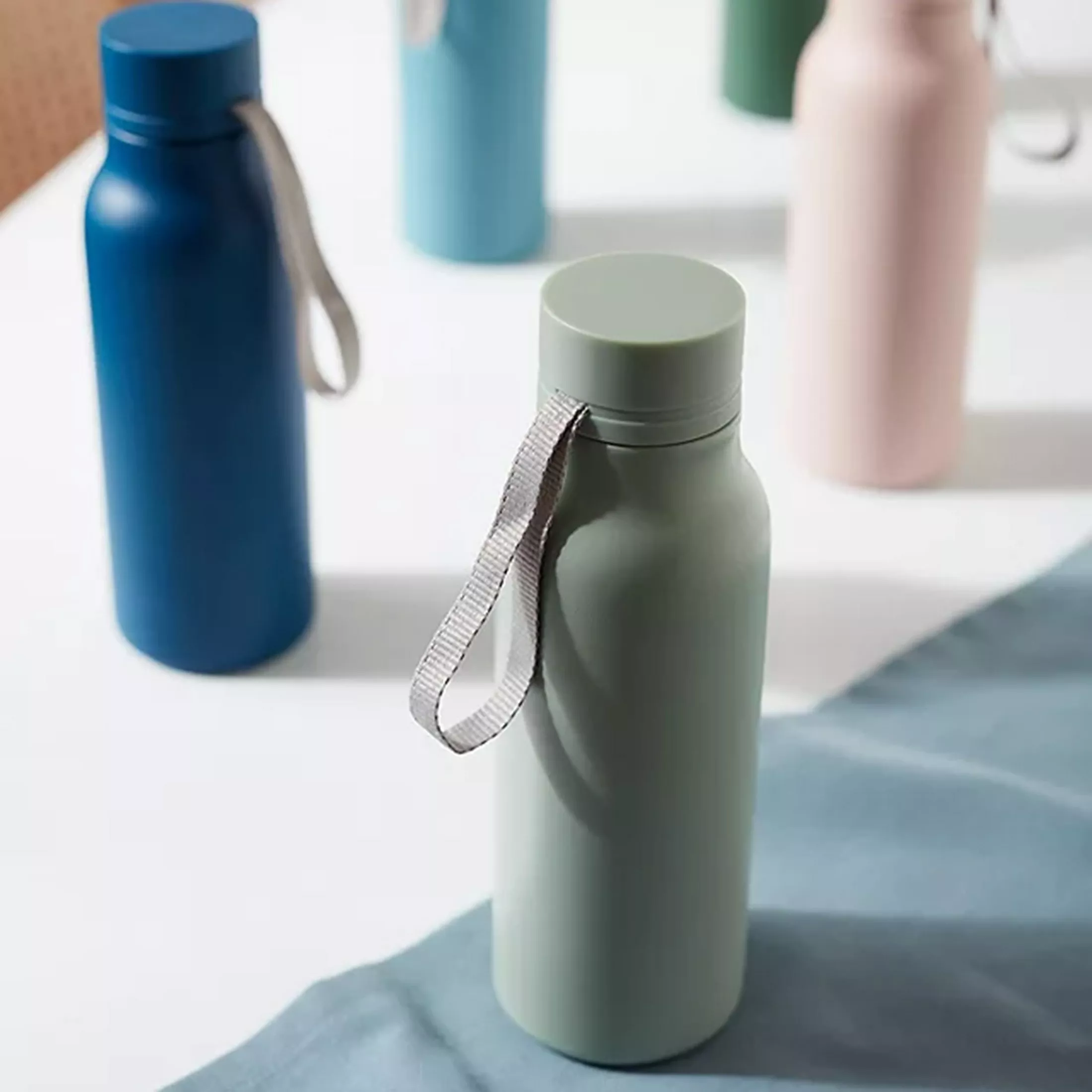 Reusable stainless steel water bottle with carry strap, in sage green, with pastel bottles blurred behind.