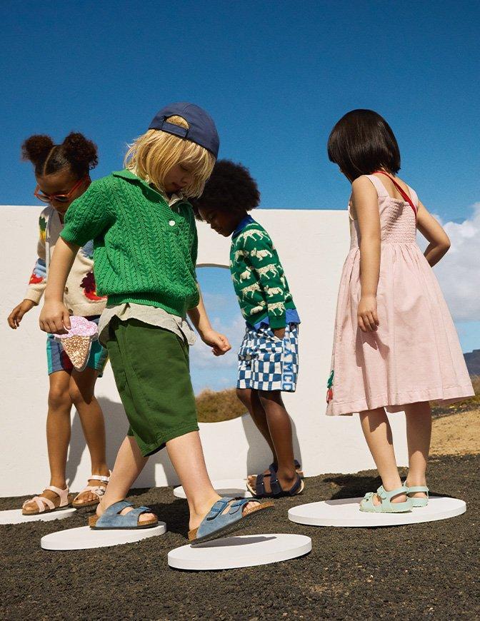 A group of four children are standing and walking on circular platforms outdoors, dressed in colorful, stylish summer clothing under a clear blue sky.