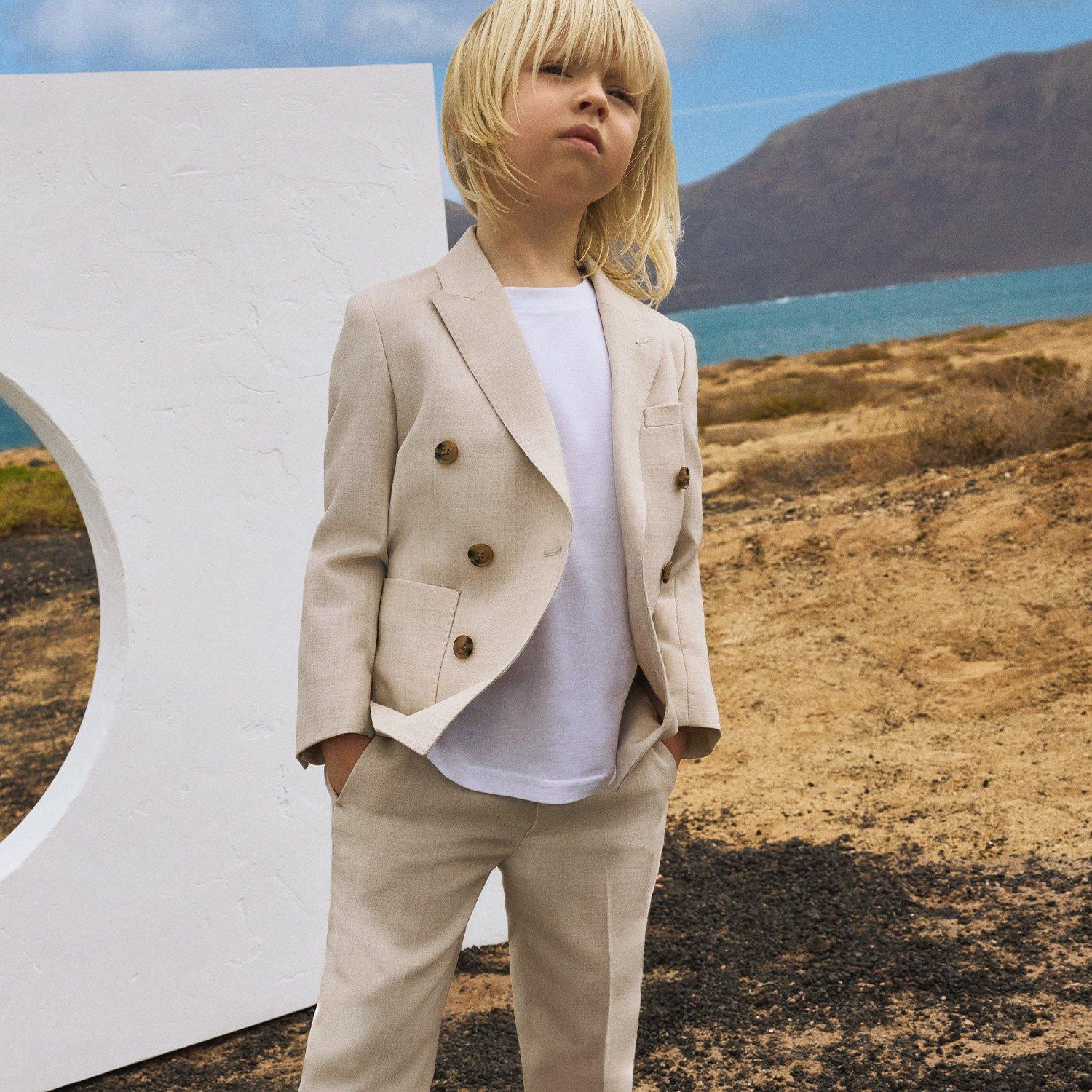Boy in cream jacket and trousers, next to white wall backdrop, lake and hillside landscape
