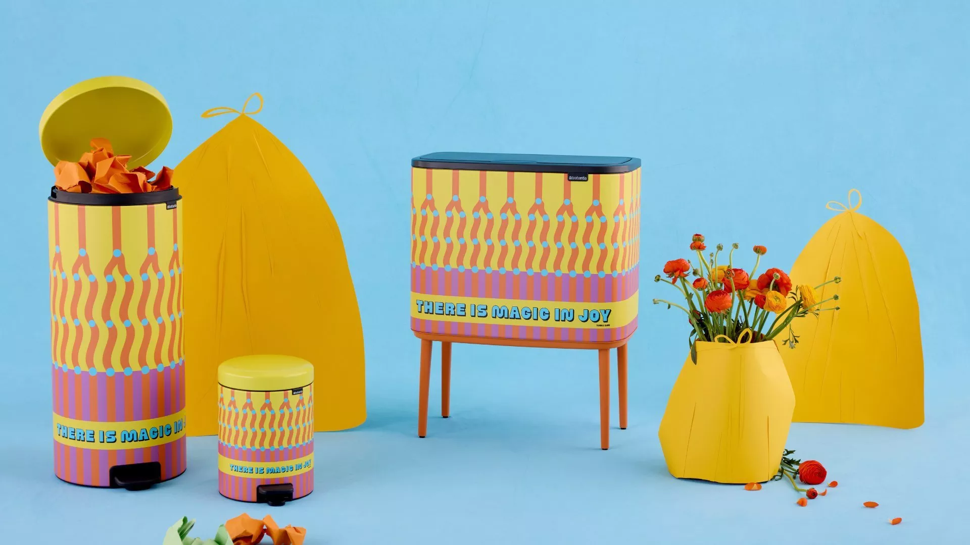 Colorful Brabantia waste bins decorated with a yellow and pink pattern reading "There is magic in joy" against a light blue background.