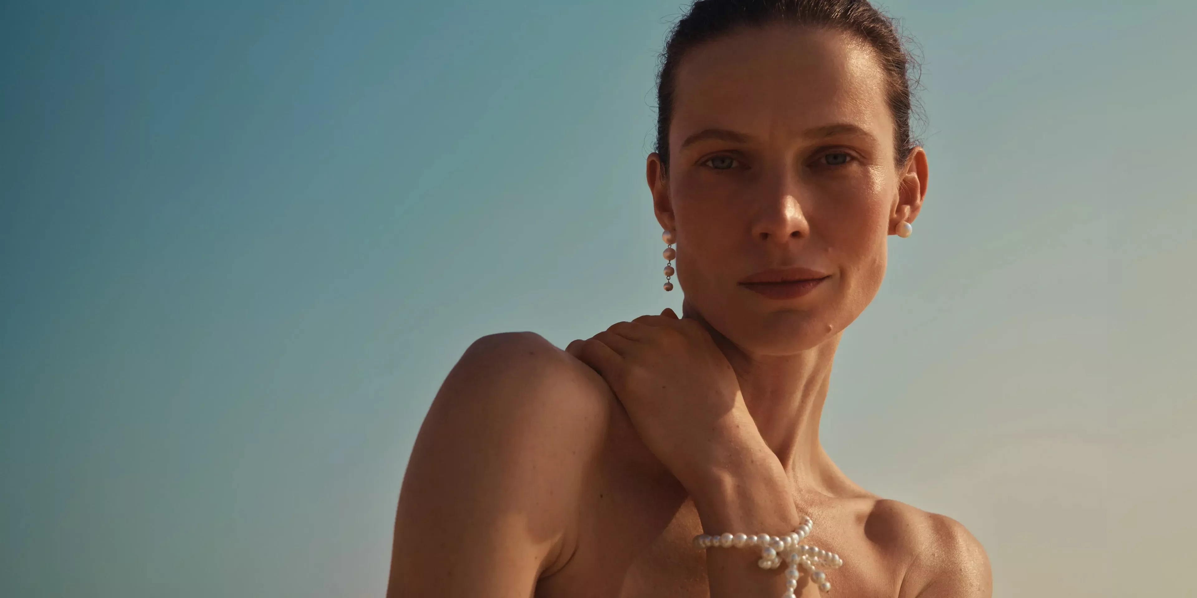 Woman wearing soft glam makeup, pearl earrings and bracelet, against clear blue sky