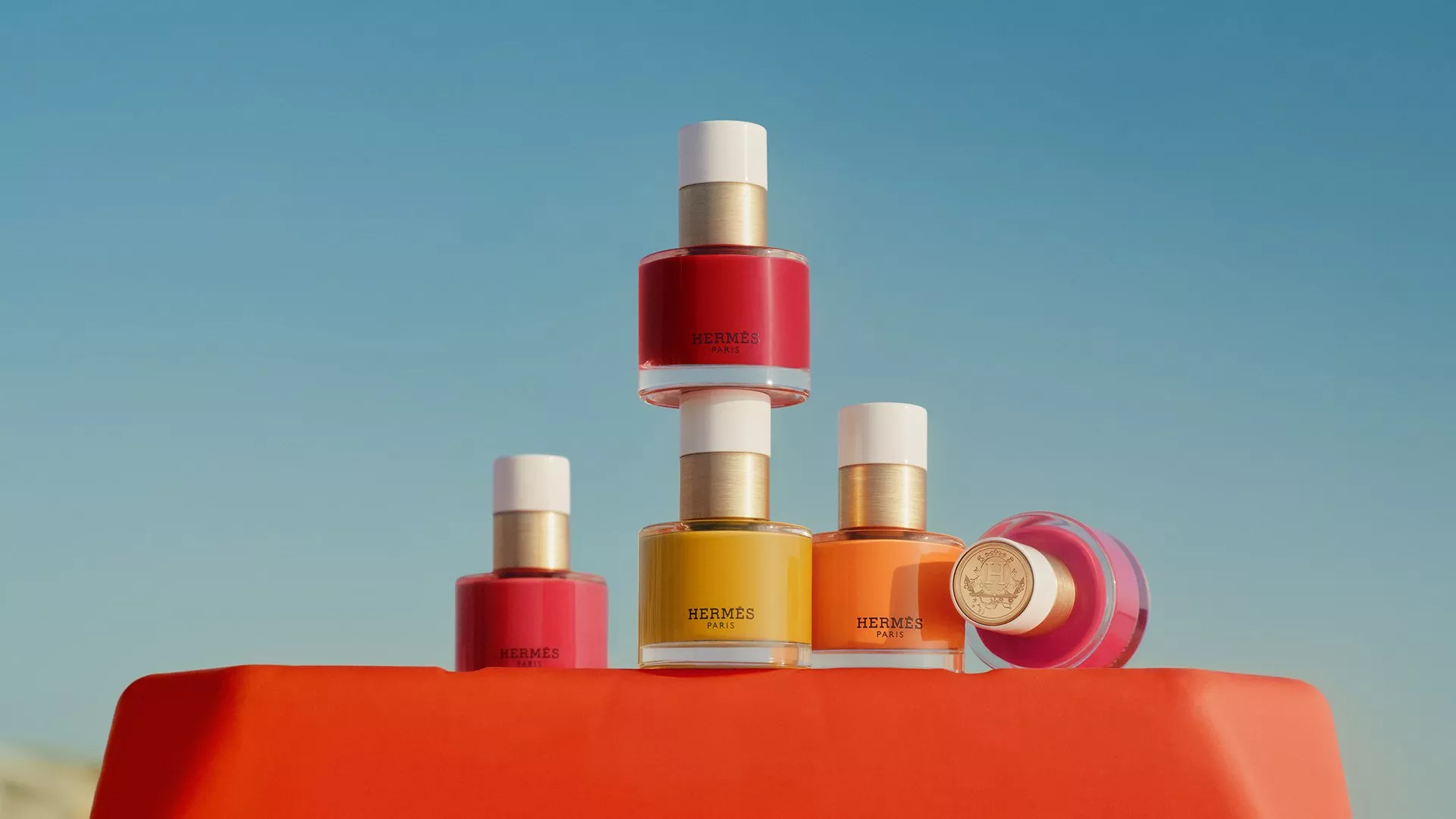 Hermès nail polish bottles in red, pink, yellow and orange arranged on a bright orange pedestal under a clear blue sky