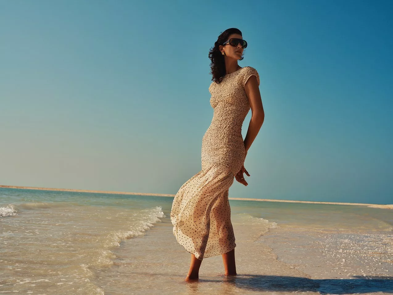 Woman in polka dot summer dress and sunglasses standing in shallow sea water on a sunny beach shoreline