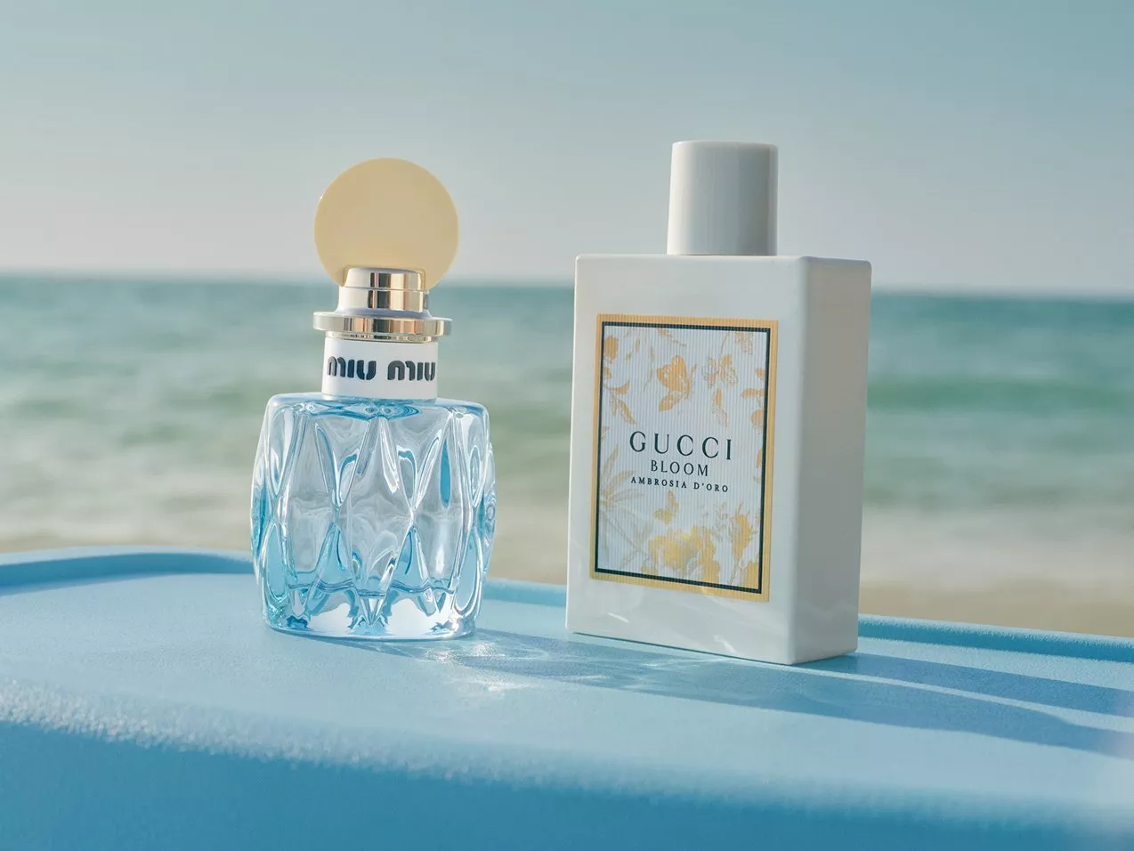 Miu Miu and Gucci Bloom perfume bottles on a blue surface by the sea, luxury fragrance product photo