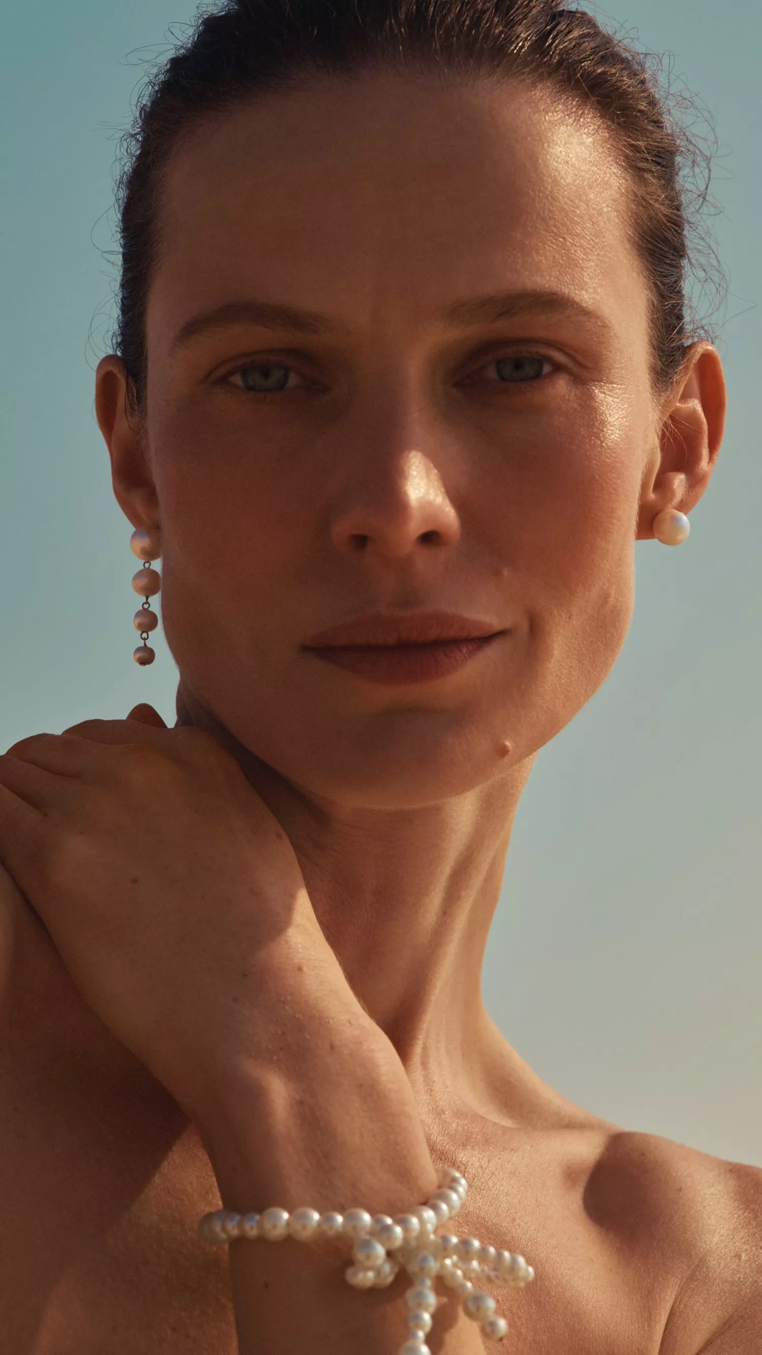 Woman wearing soft glam makeup, pearl earrings and bracelet, against clear blue sky