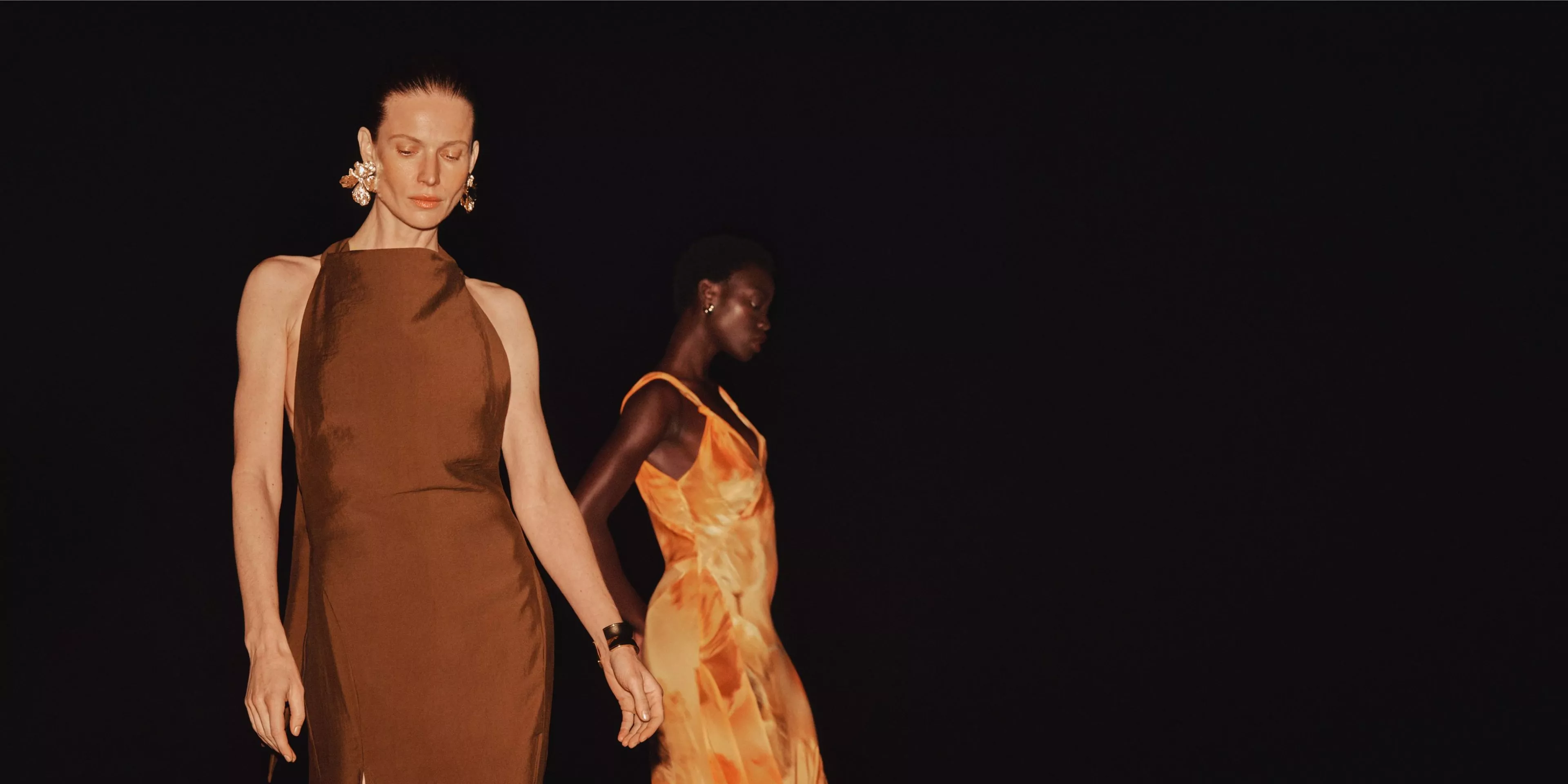 Fashion runway models wearing elegant brown and orange satin dresses on a dark catwalk, eveningwear couture show
