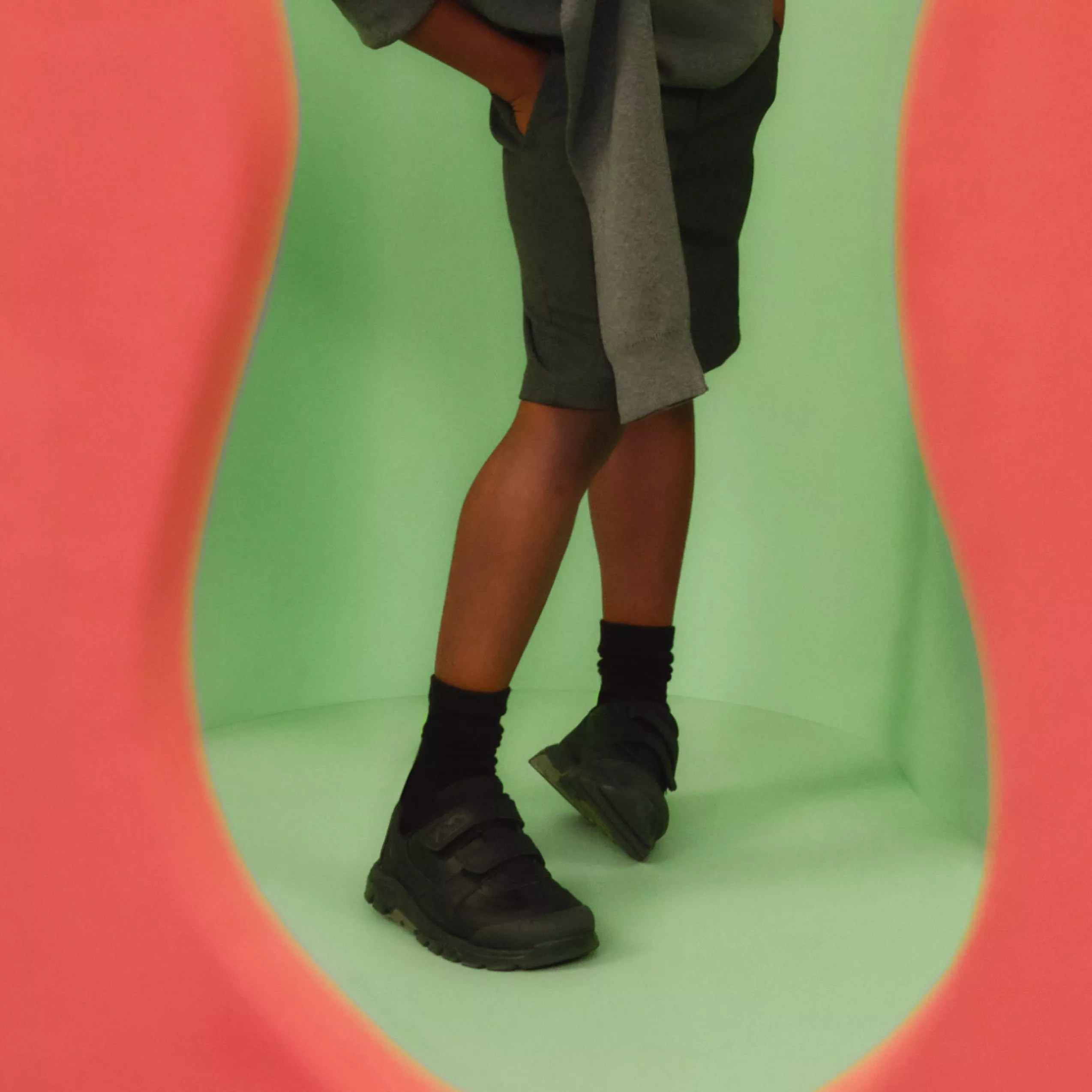 Studio fashion shot of model legs in black chunky trainers and socks, against a green backdrop with pink edges