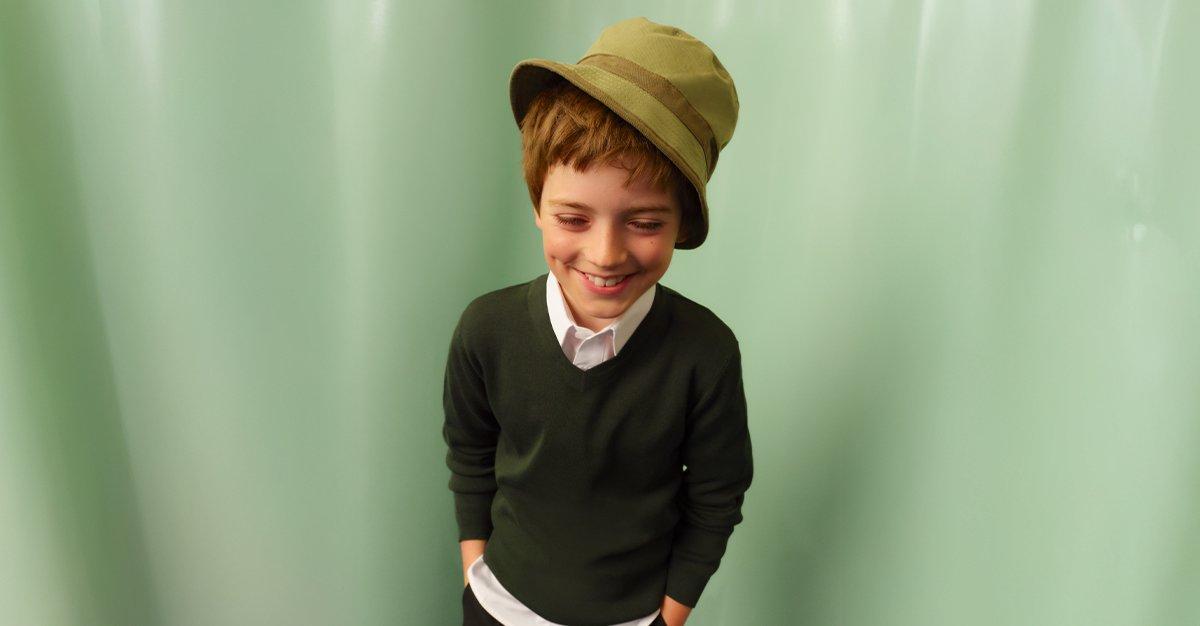 Smiling child in a green sweater and hat with hands in pockets.
