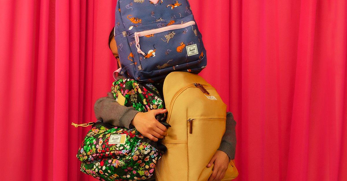 Child holding multiple colorful backpacks in front of a red curtain.