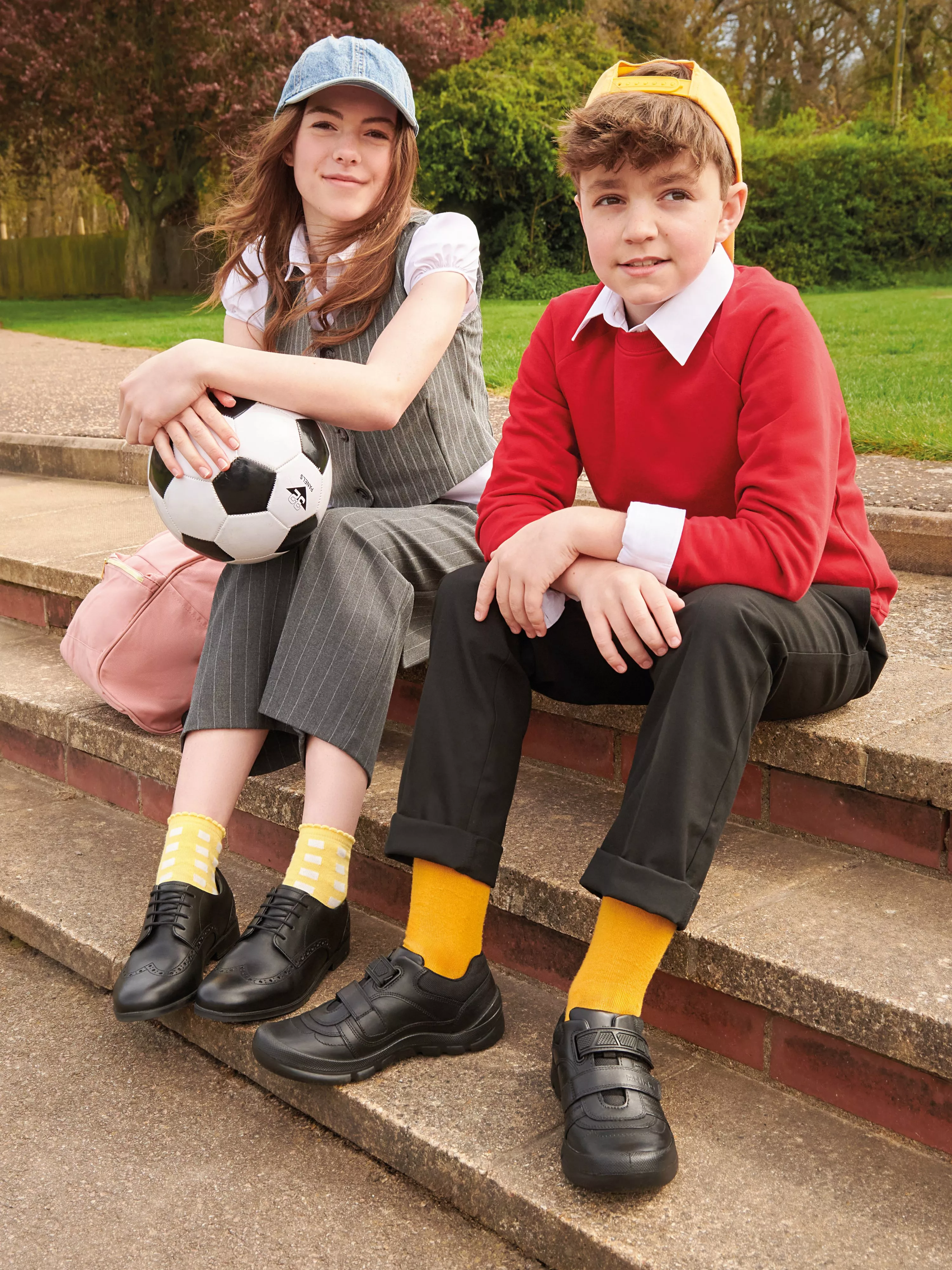 Two schoolchildren in uniform sit on outdoor steps with a football, wearing caps and bright yellow socks.