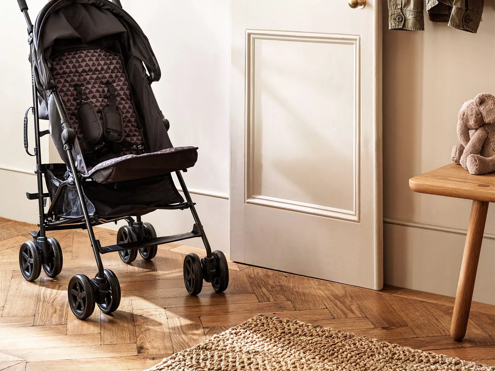 A stylish black pram with geometric patterns stands on a wooden floor, next to a plush toy on a stool.
