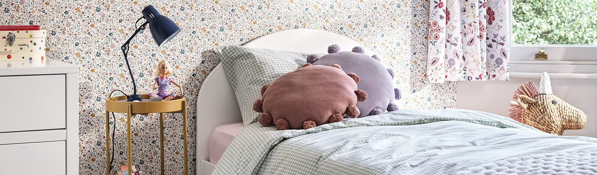 Cosy children’s bedroom with floral wallpaper, white bed, gingham bedding, pom-pom cushions and bedside lamp
