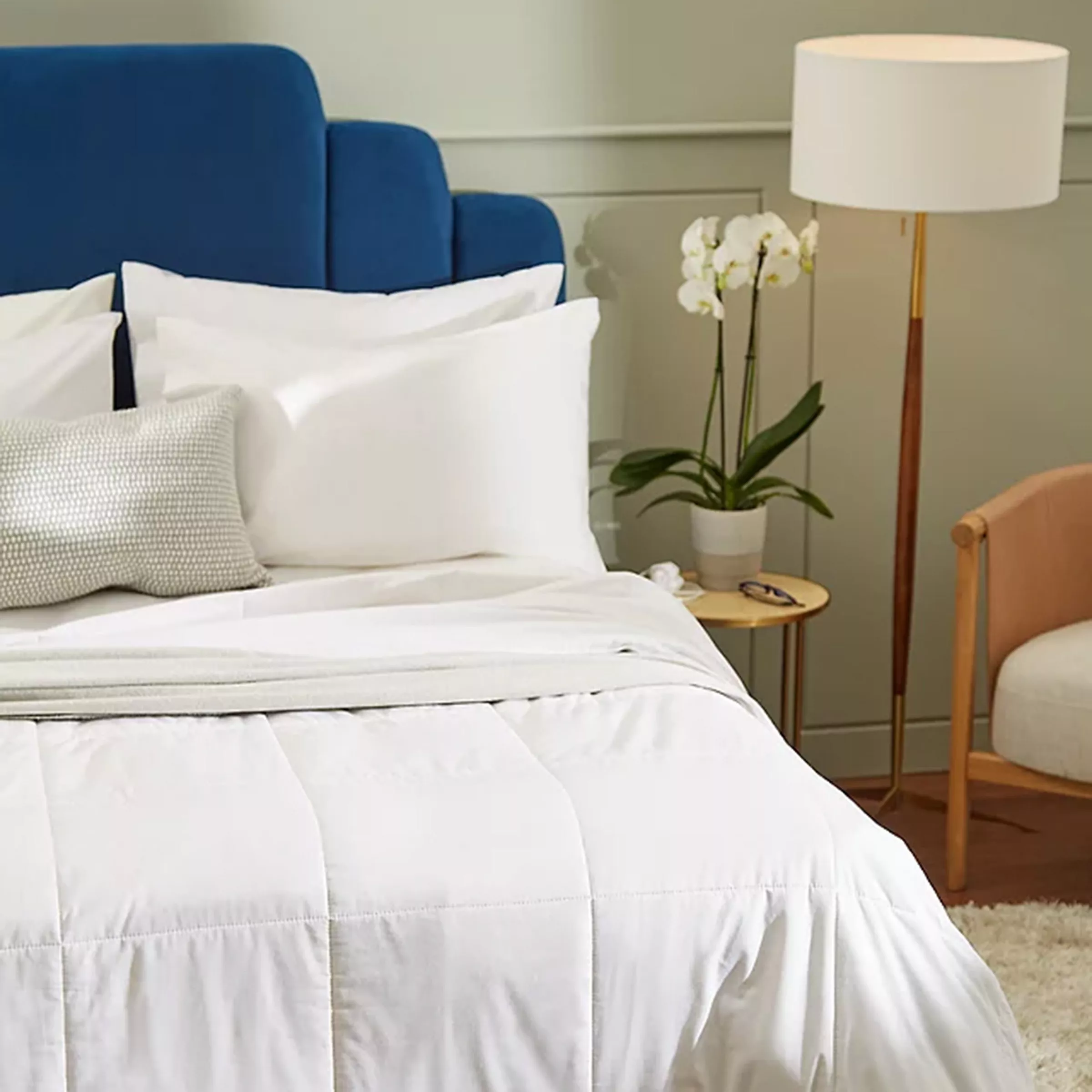 Elegant bedroom with blue headboard, white bedding, cosy chair, and side table with orchid plant and lamp.