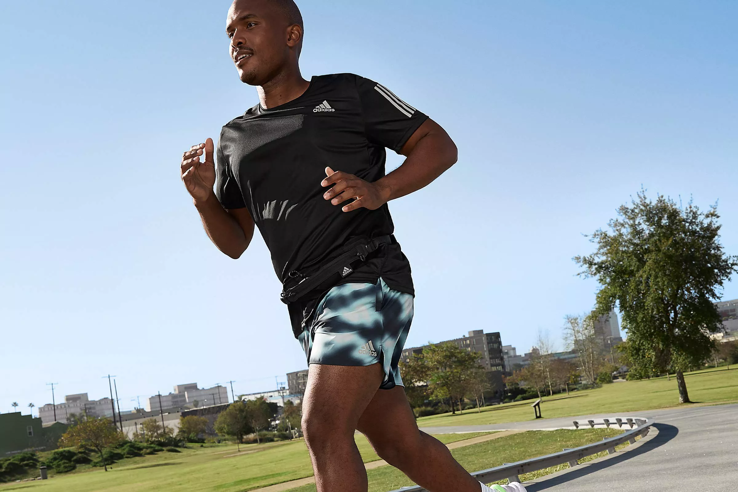 Male runner jogging outdoors in a park on a sunny day, wearing black Adidas sportswear and running shorts