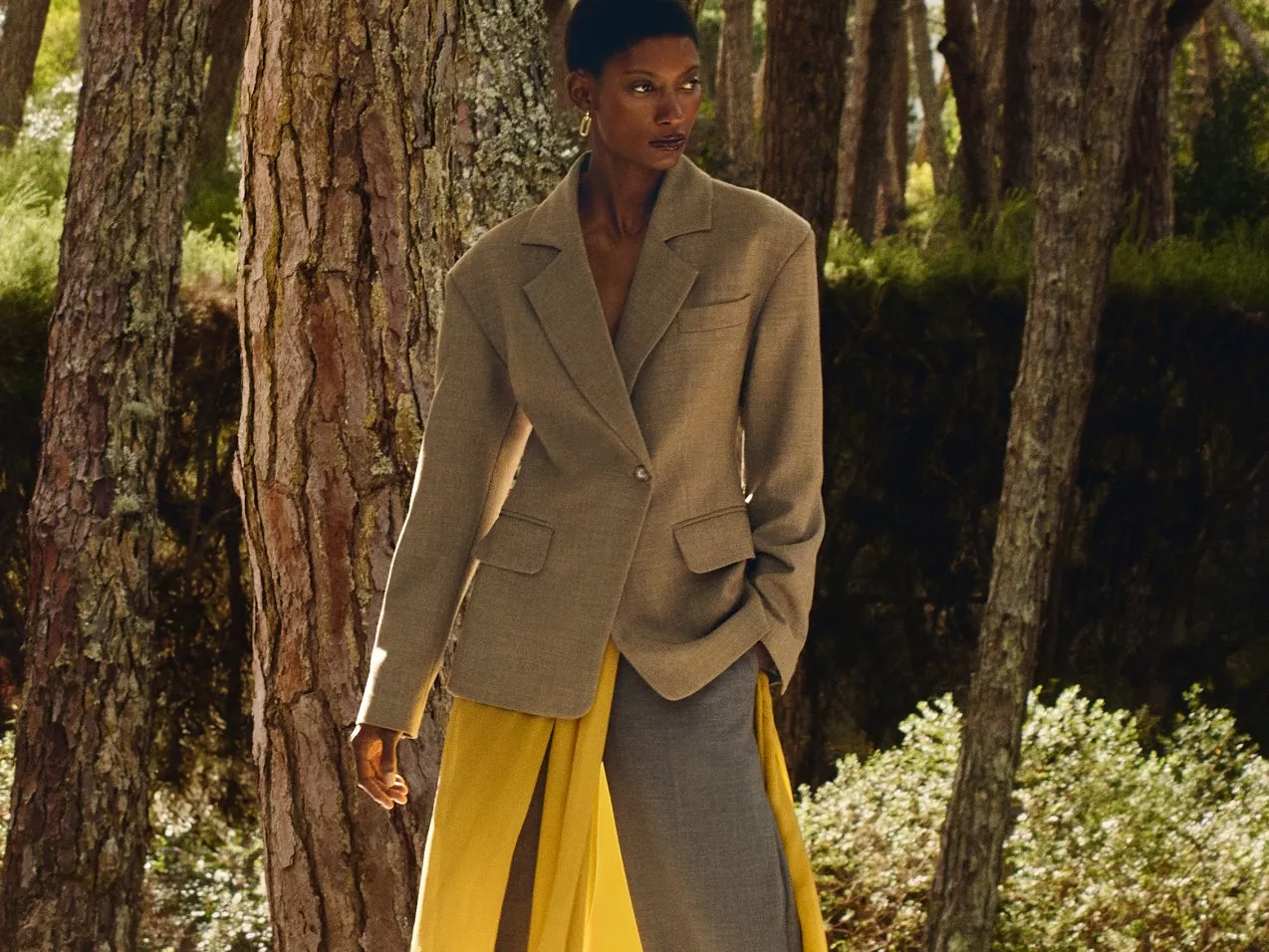 Stylish outfit featuring a beige blazer and flowing yellow skirt in a serene woodland setting.