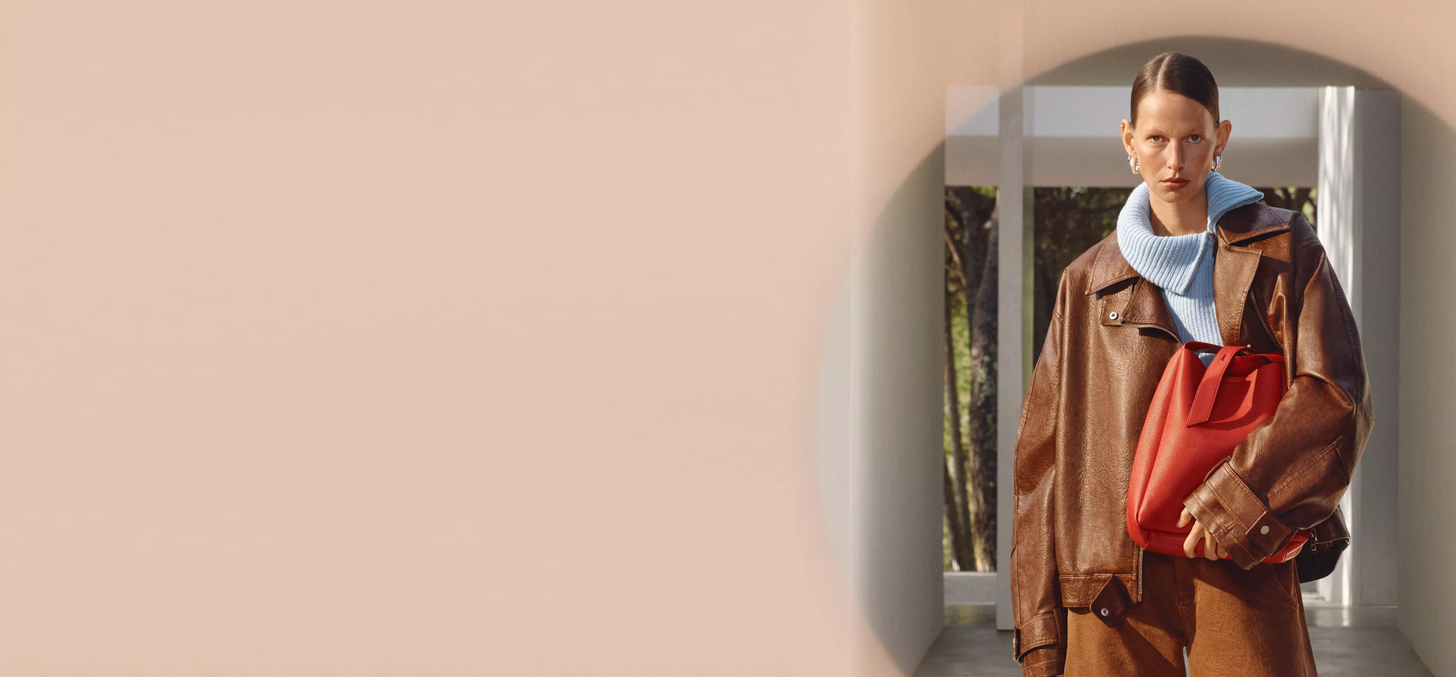 A stylish person in a brown leather outfit holds a vibrant red handbag, with a modern architectural backdrop.