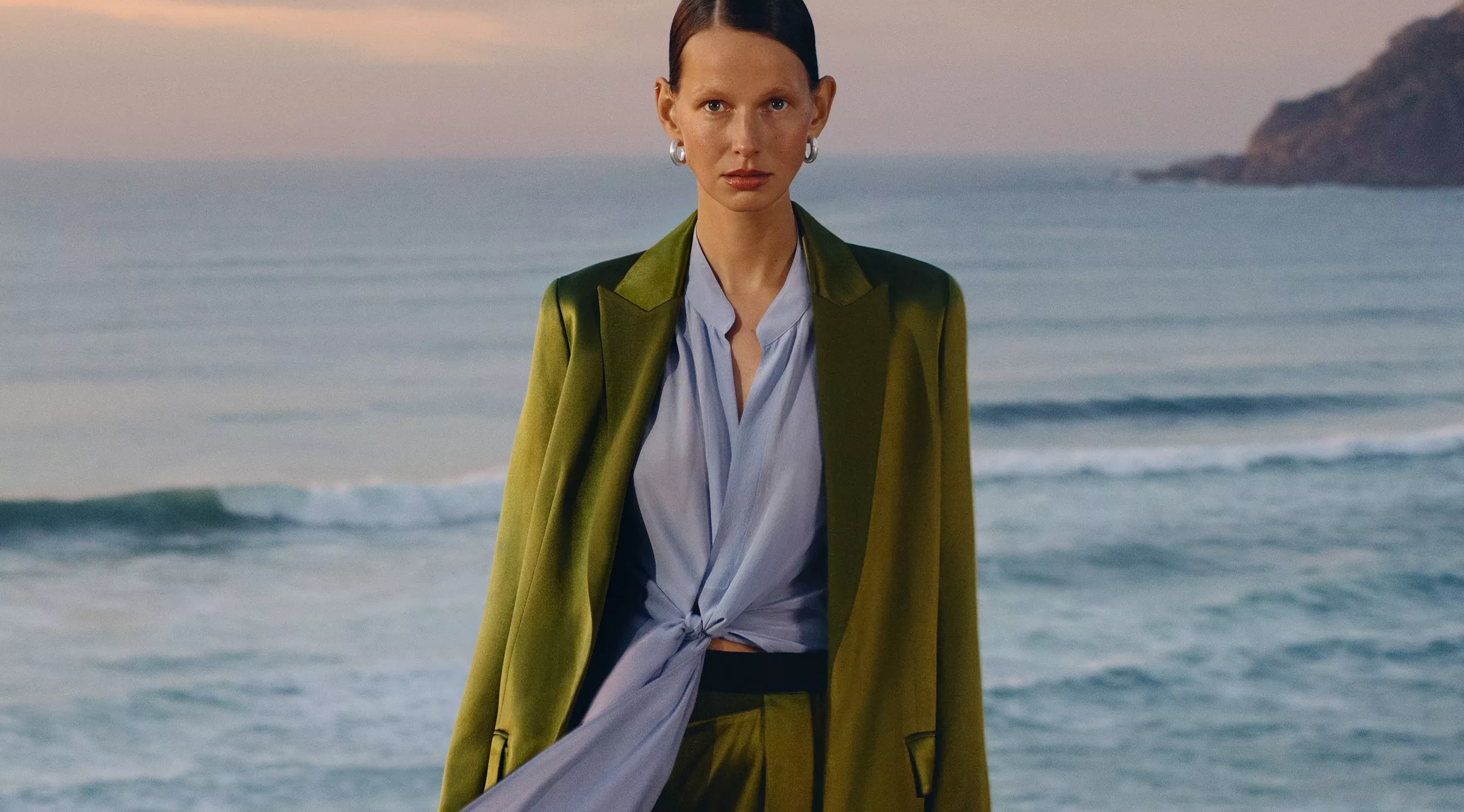 A stylish outfit featuring an olive green blazer over a light blue blouse, set against a scenic ocean backdrop.