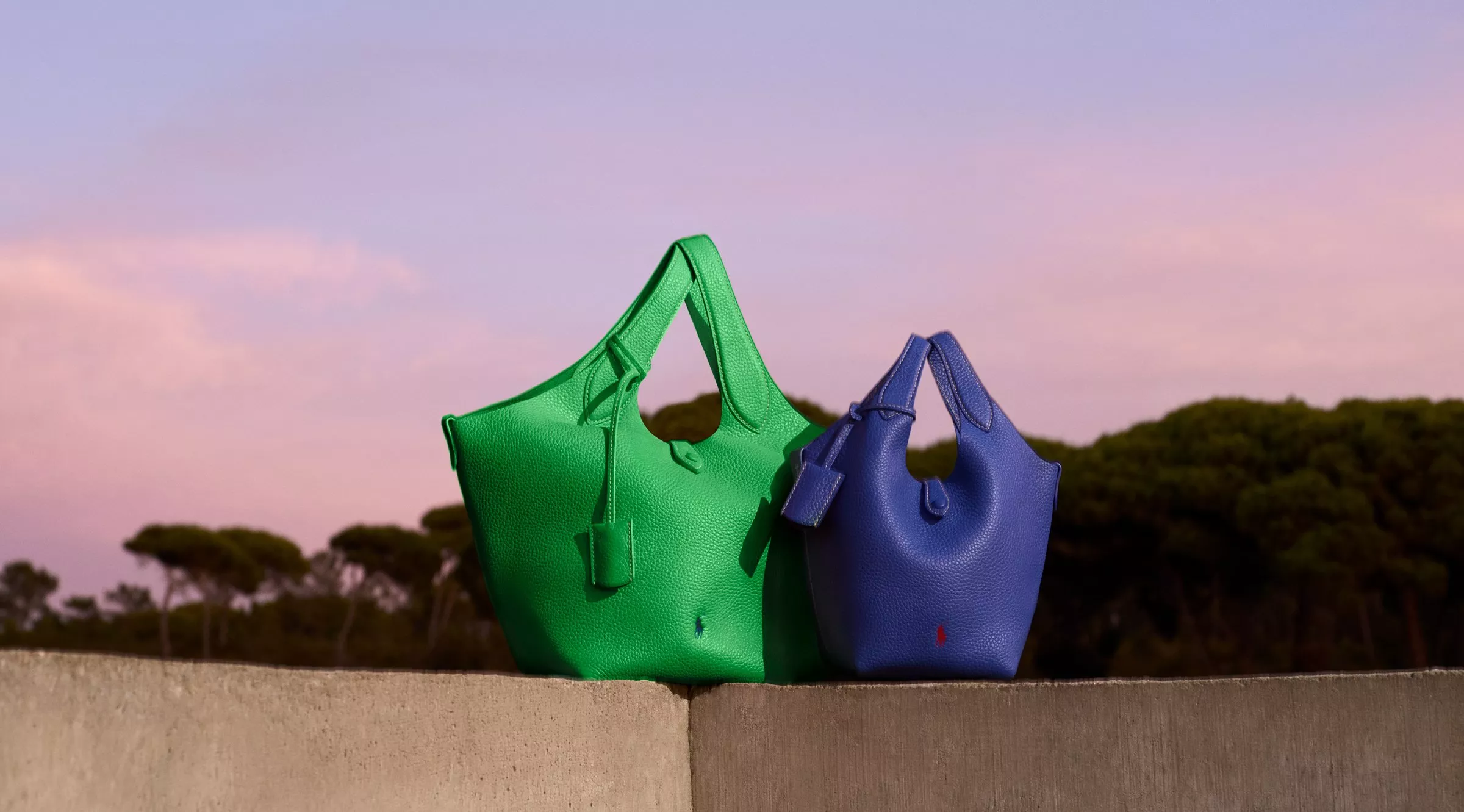 Two stylish Ralph Lauren leather handbags, one green and one blue, displayed against a serene pink and purple sunset sky.