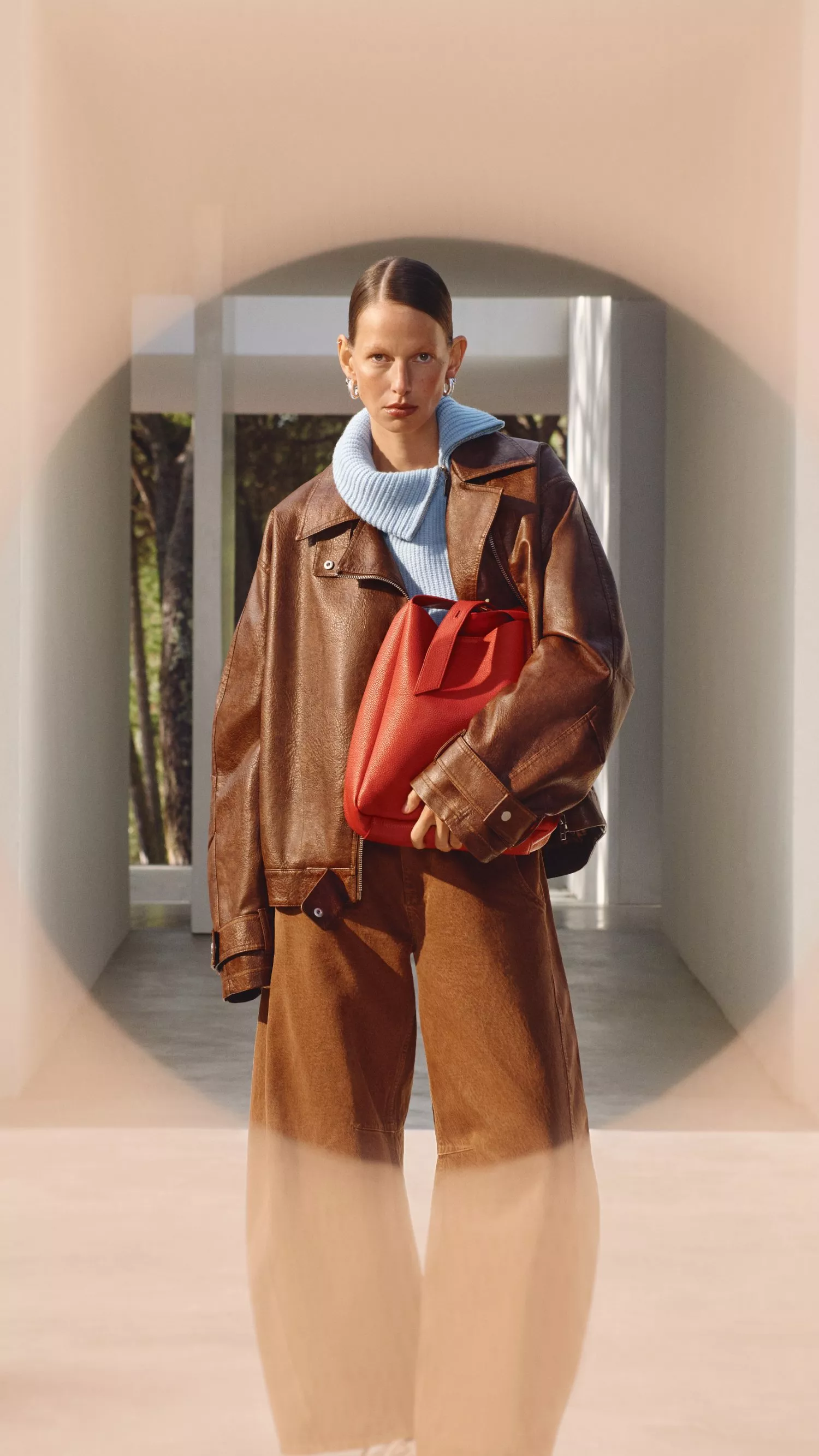Model in brown leather outfit and light blue sweater, holding a red handbag, stands in a modern interior setting.