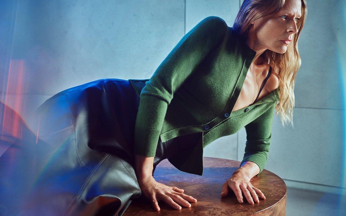 Female model sat on a table wearing a green cashmere cardigan and black leather midi skirt