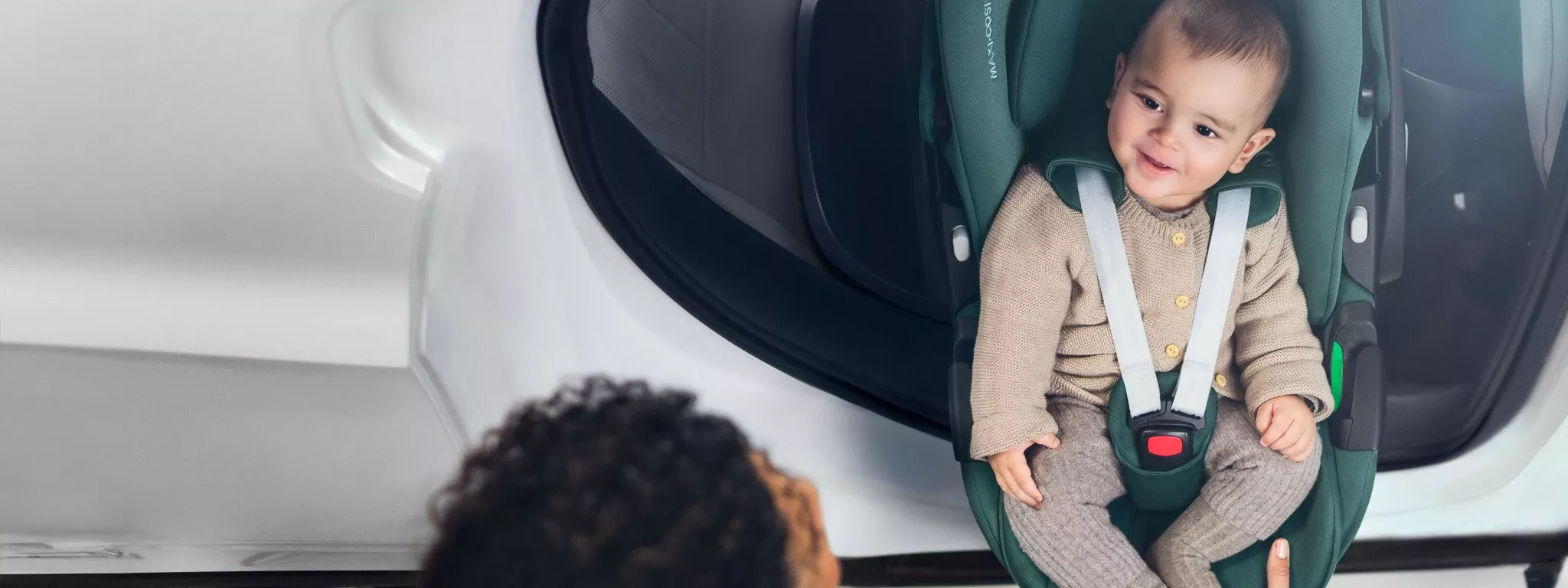 A baby in a green car seat with an adult nearby. The car door is open, showing a white vehicle interior.
