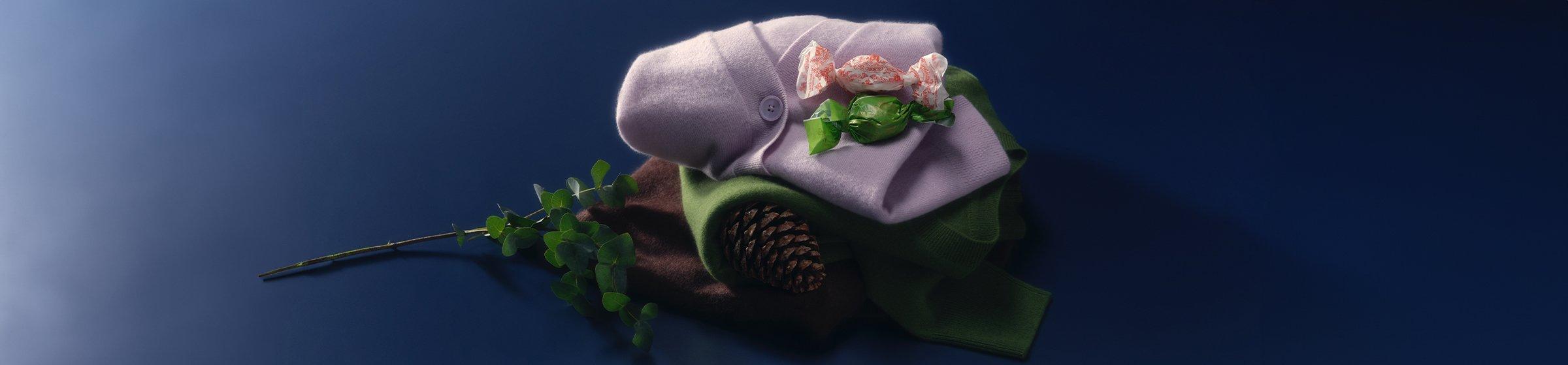 A pile of cashmere jumpers and cardigans on a navy backdrop next to a pine cone and christmas sweets