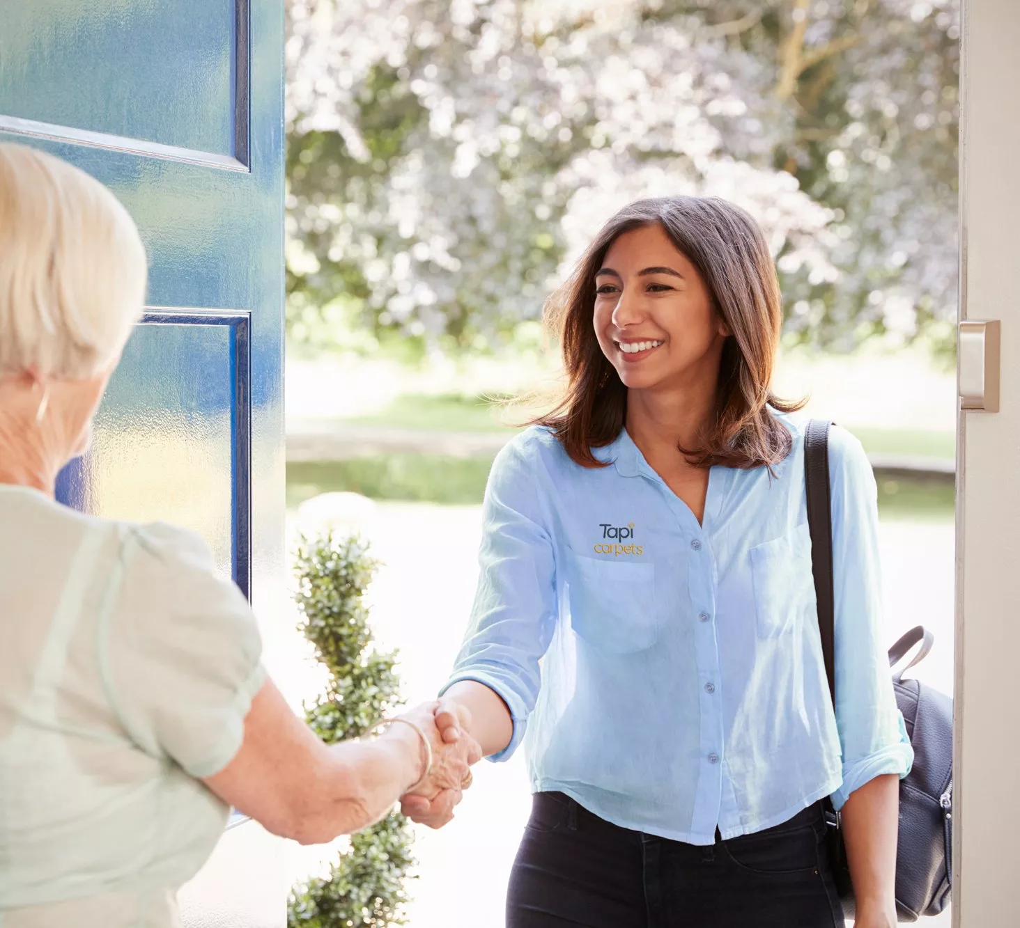 A person wearing a "Tapi Carpets" shirt shakes hands with a woman at a doorway, with a lush garden visible outside.