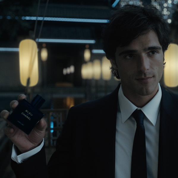 Man in dark suit holding Bleu de Chanel perfume bottle in a stylish, dimly lit modern interior setting