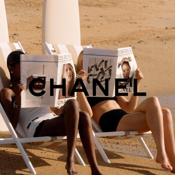 Two models sunbathe on beach loungers, reading Les Beiges newspapers in a Chanel summer beauty campaign.