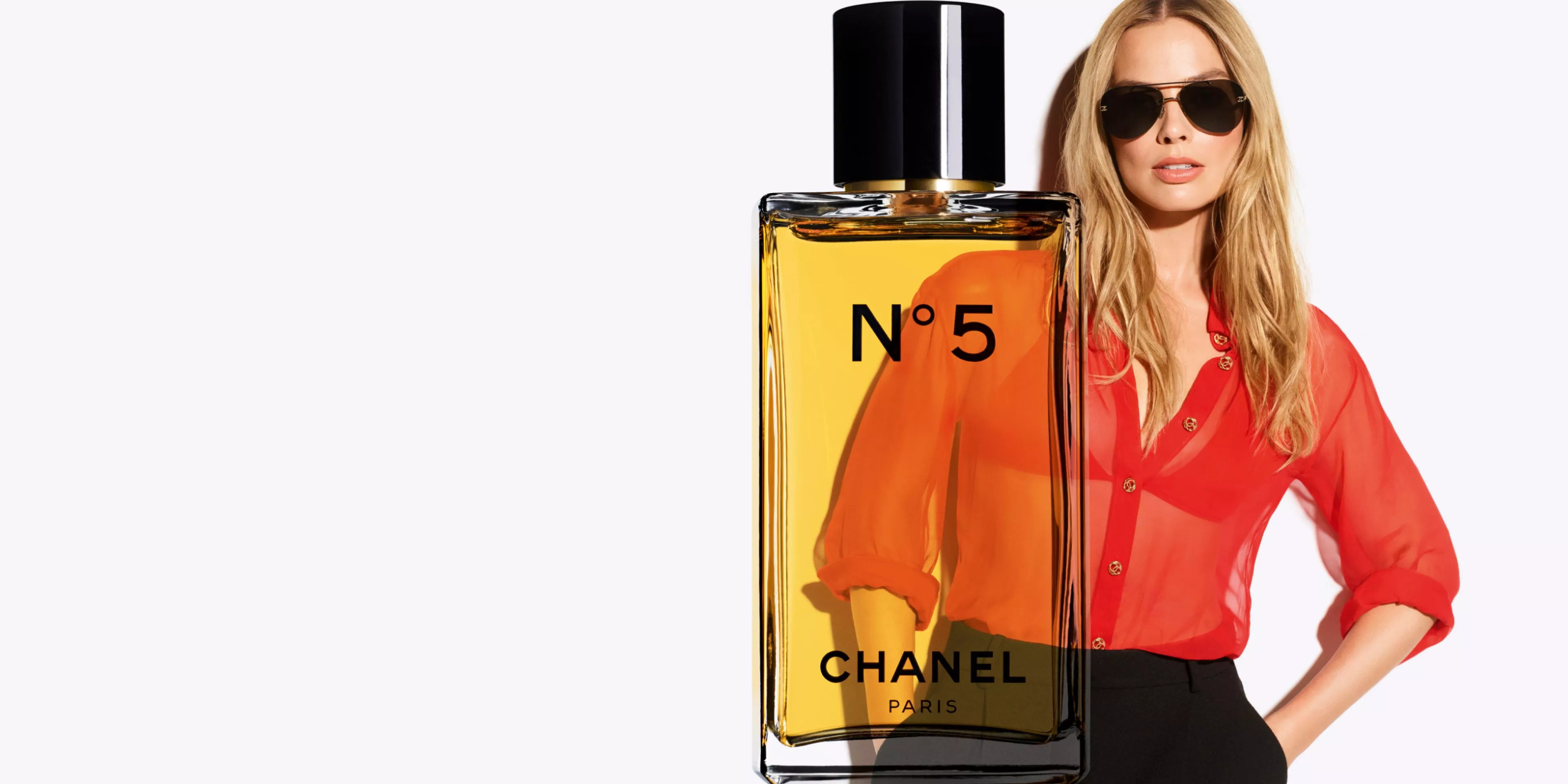 Chanel No. 5 perfume bottle with black cap, alongside a person in a sheer red blouse, on a light background.