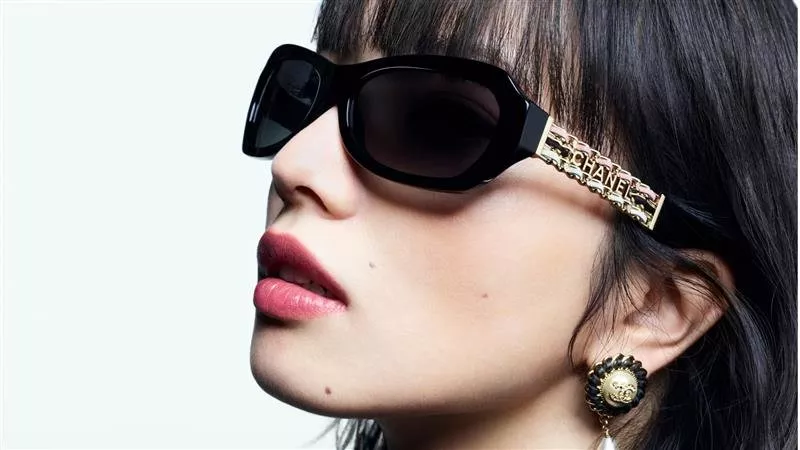A close-up profile of a person wearing oversized black rectangular Chanel sunglasses with ornate gold lettering on the temples