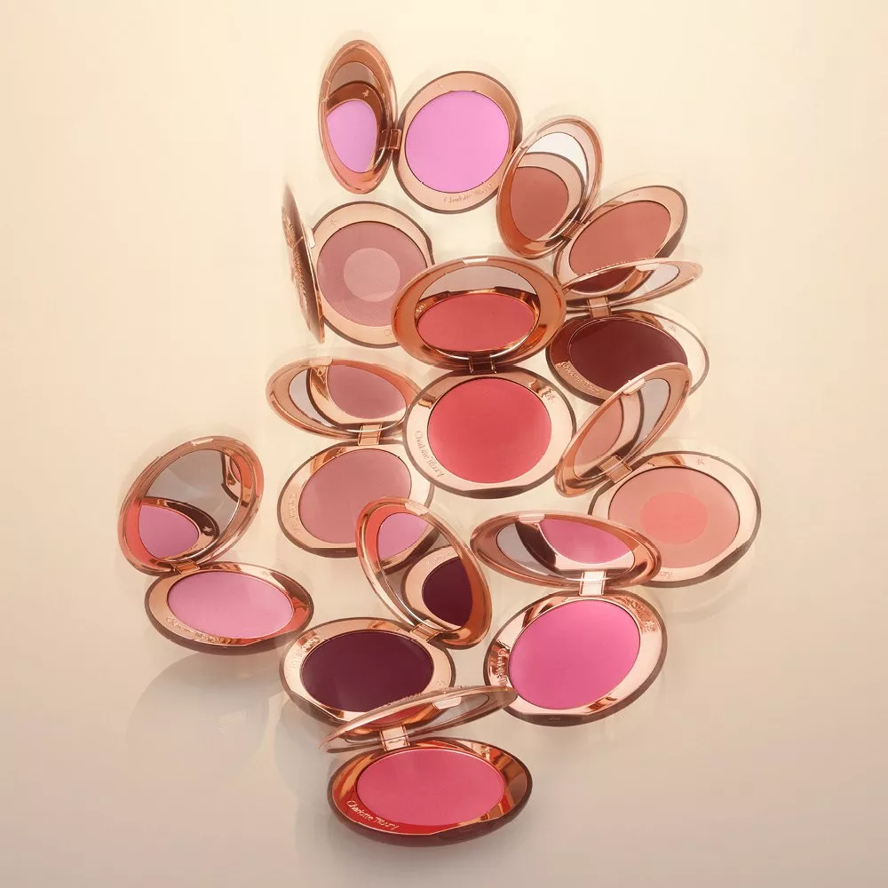 Stacked cream blush and highlighter sticks in rose-gold tubes on a pink background, glossy beauty product shot