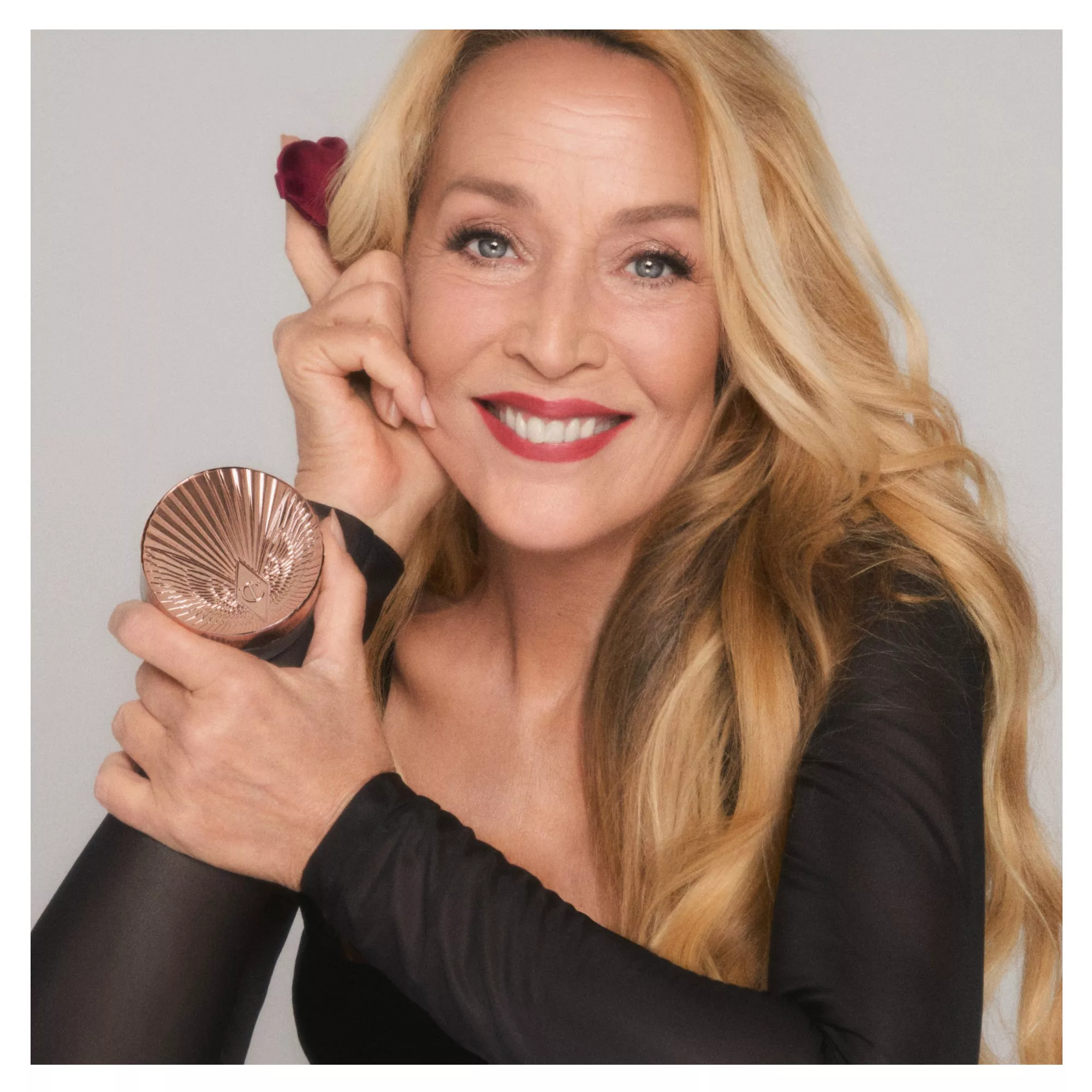 Smiling blonde woman holding a rose-gold makeup compact, showcasing red lipstick and glowing skin in beauty portrait.