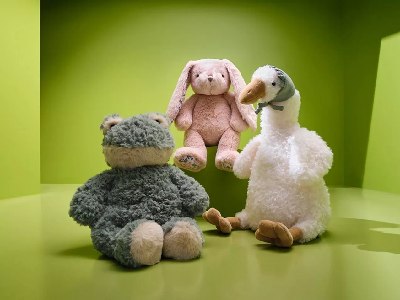 Three soft plush toys: grey teddy bear, pink bunny and white goose on a bright green background, nursery decor photo