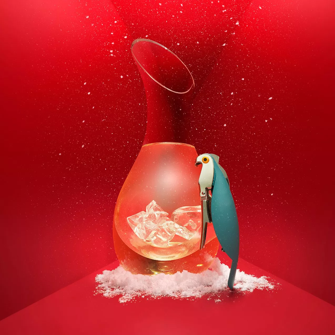 Glass carafe with ice cubes and snow, beside a blue parrot-shaped corkscrew on festive red background