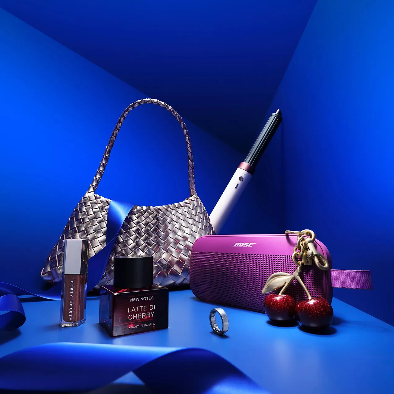 Luxury still life with woven silver handbag, Bose purple speaker, cherry charm, perfume and lipstick on blue backdrop