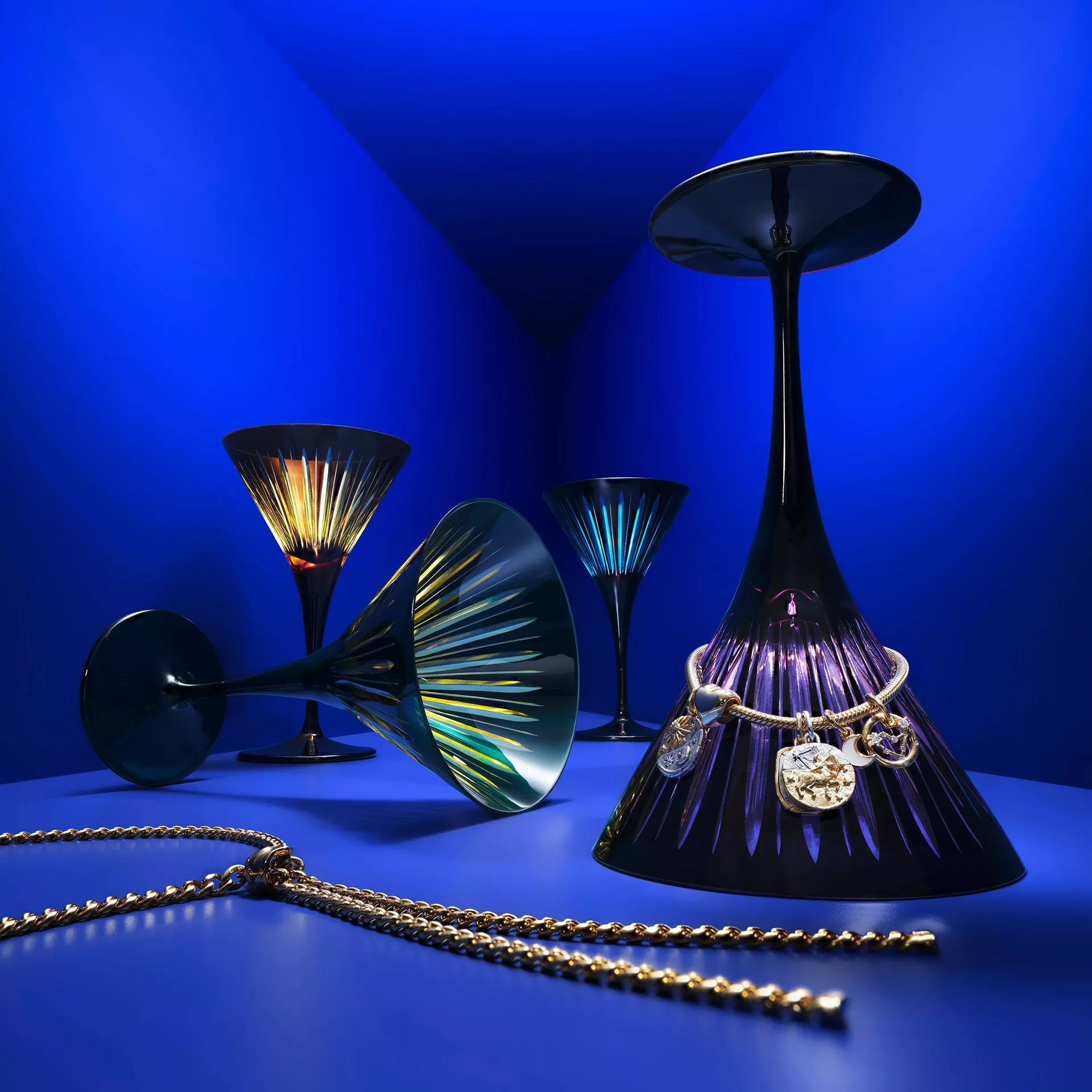 Luxury coloured crystal cocktail glasses and gold charm jewellery on a vivid blue studio background