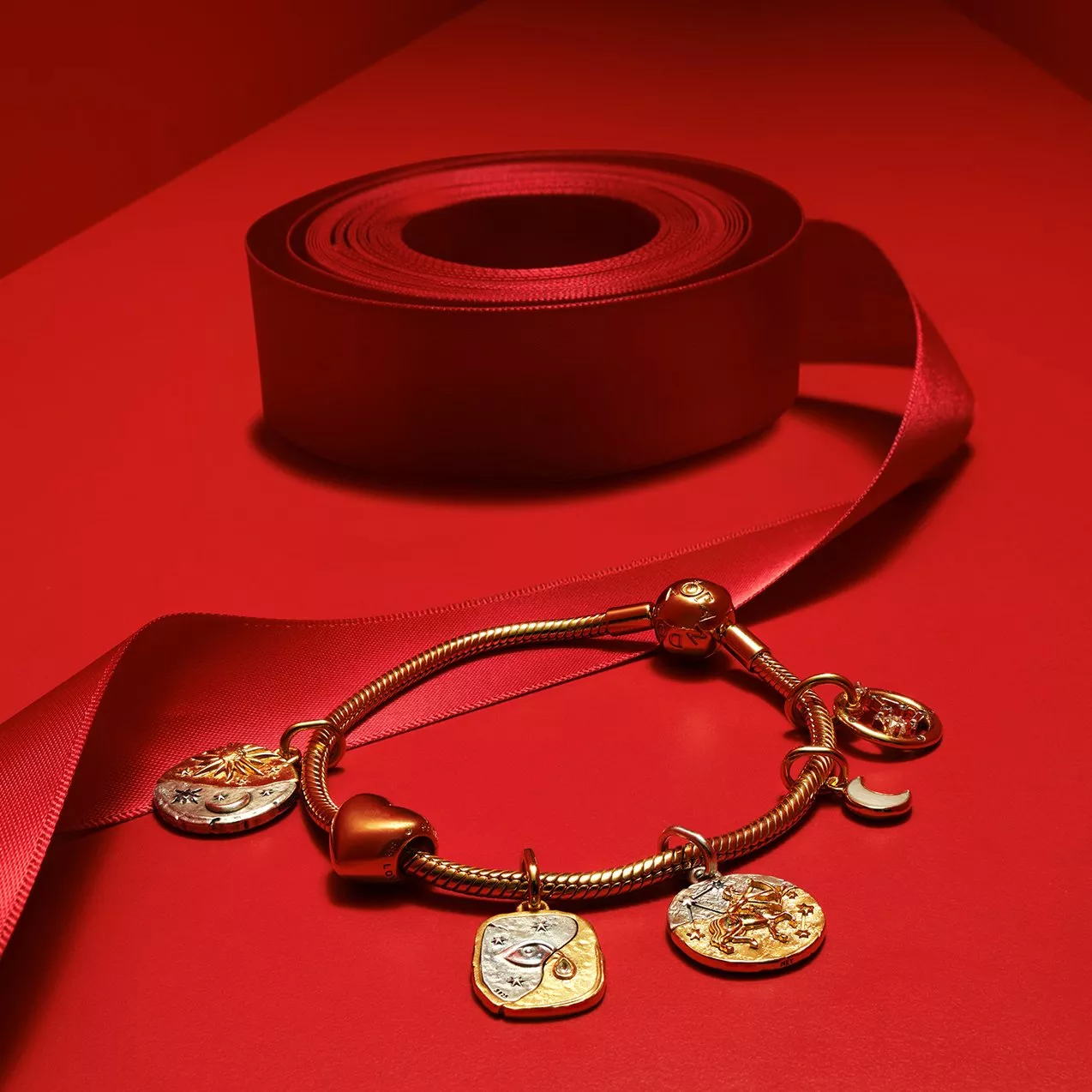 Gold charm bracelet on red backdrop with rolled satin ribbon, luxury jewellery still life close-up