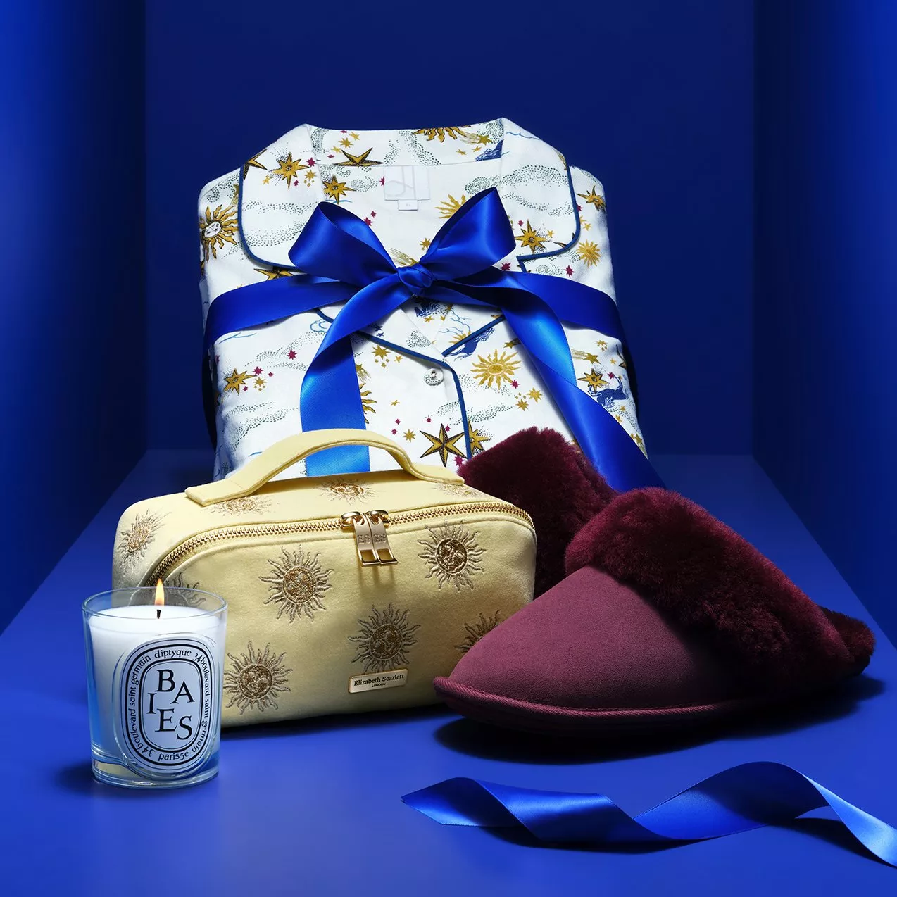 Gift set with star-print pyjamas tied with blue ribbon, candle, embroidered bag and burgundy slippers on blue background