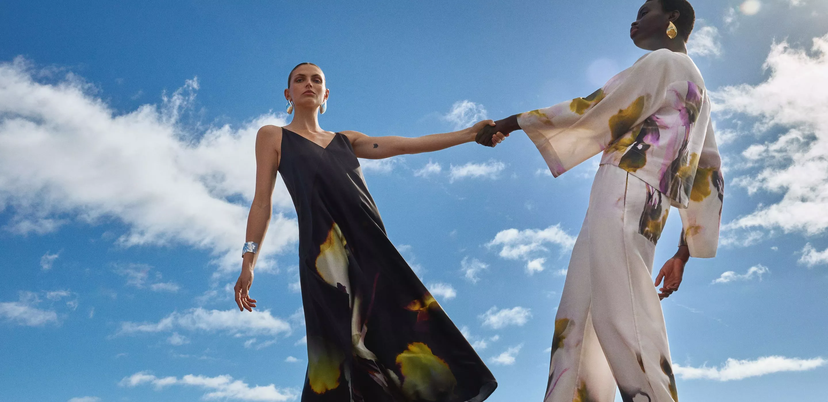 Two fashion models holding hands in printed dresses and trousers, shot from below against a blue sky with clouds
