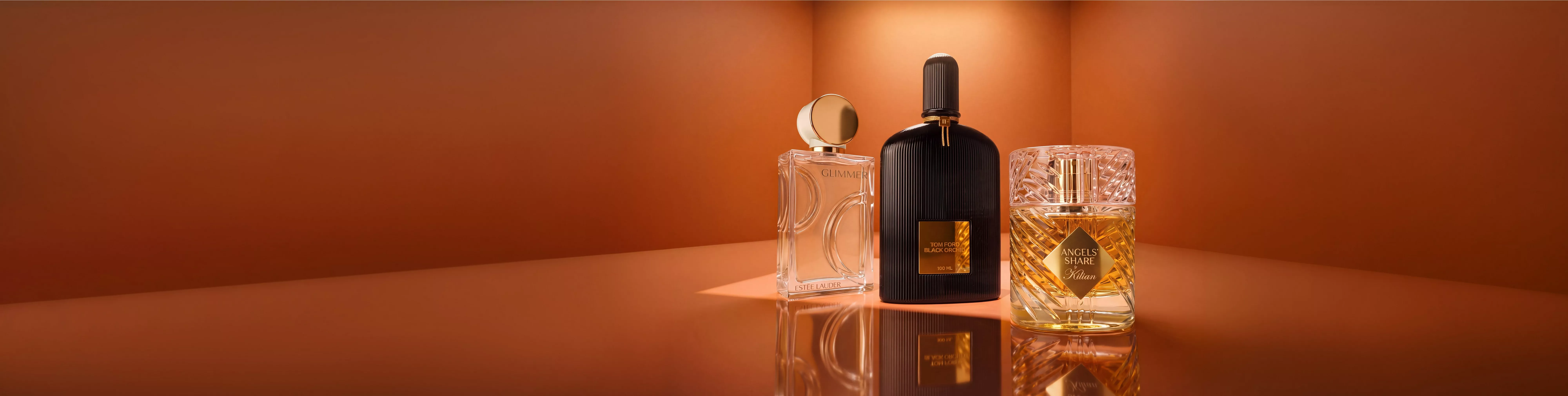 Three luxury perfume bottles, including a black Tom Ford fragrance, displayed on a reflective surface against a warm brown background.