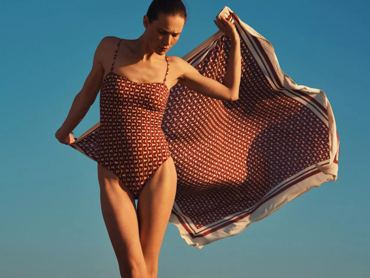 Woman in patterned one-piece swimsuit holding flowing beach scarf against clear blue sky in warm sunset light