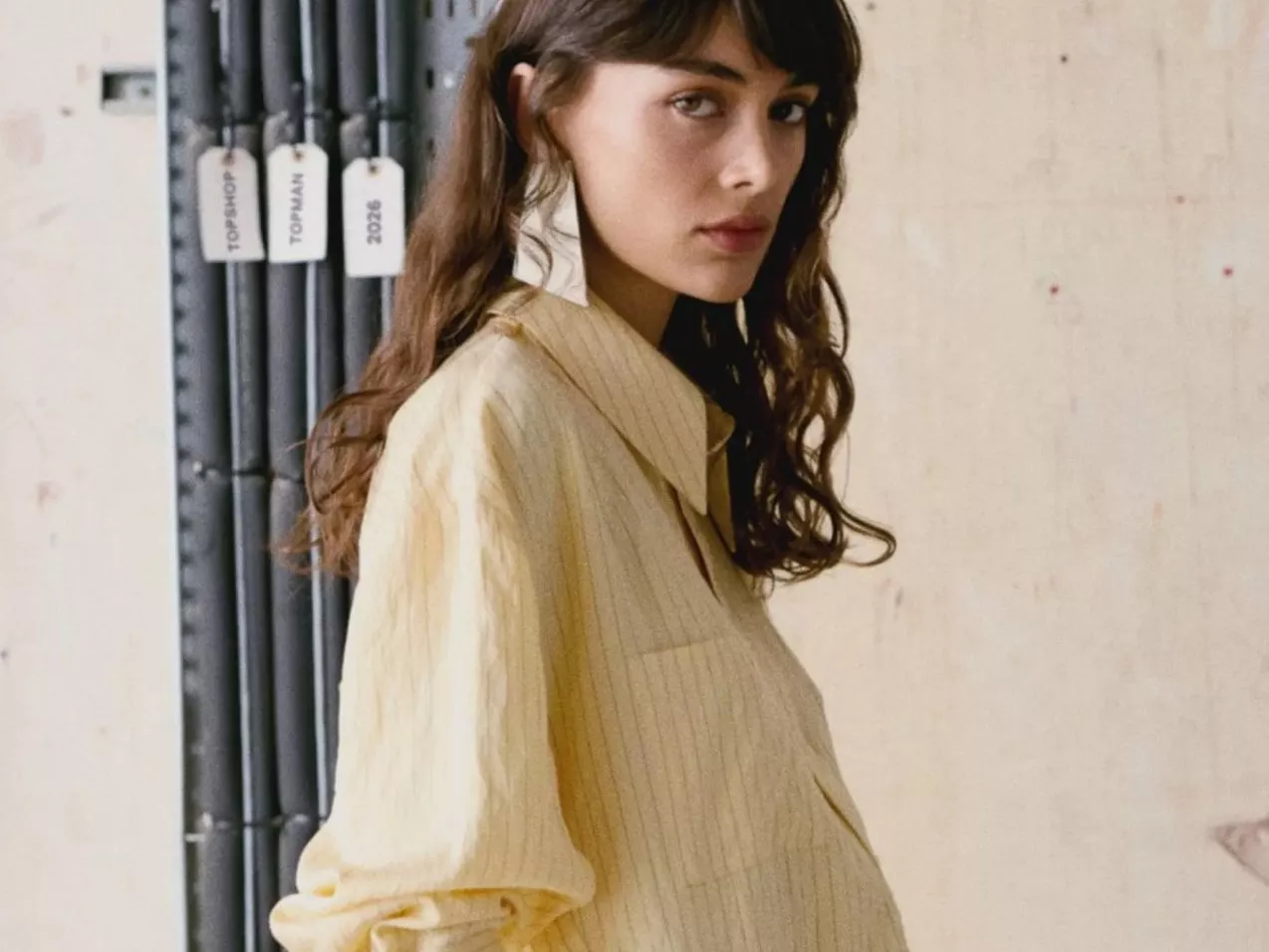 Fashion portrait of a woman in a cream striped shirt, side profile indoors with industrial pipes behind.