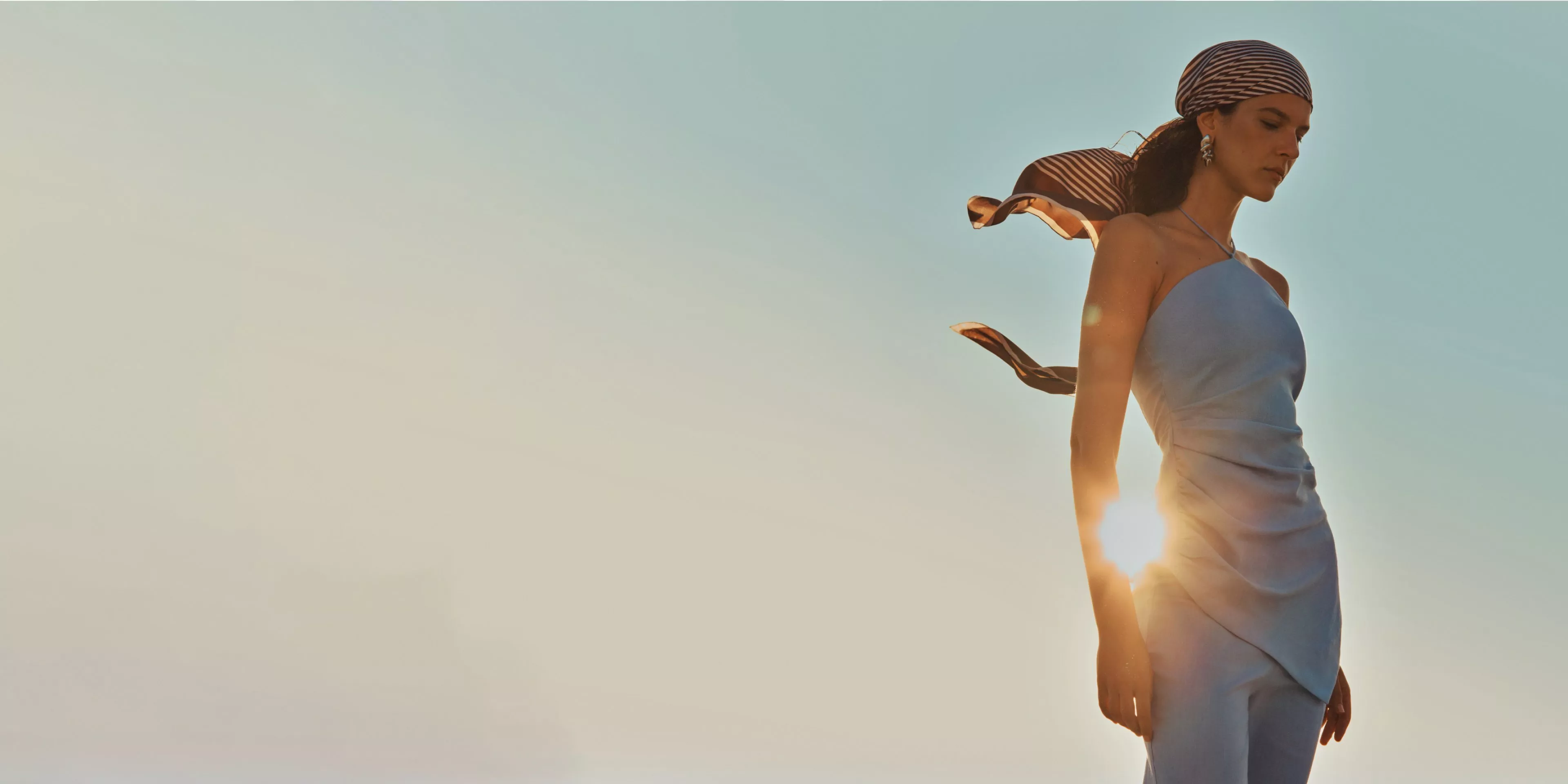 Fashion model in strapless satin dress with striped headscarf, backlit by sunset against clear sky