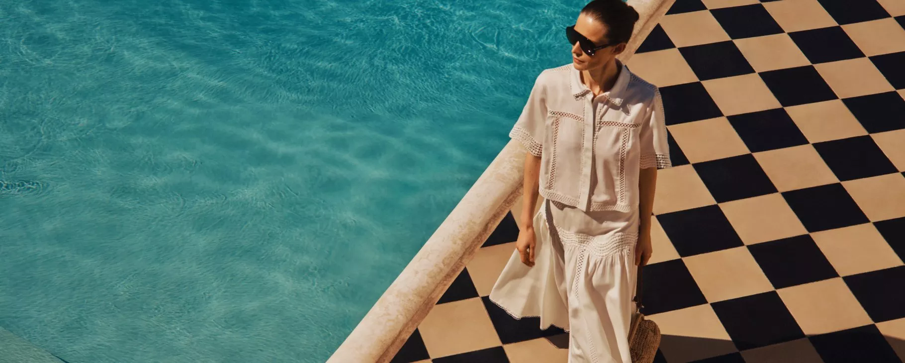 Woman in white summer outfit and sunglasses walking beside a turquoise pool on a black-and-cream tiled terrace