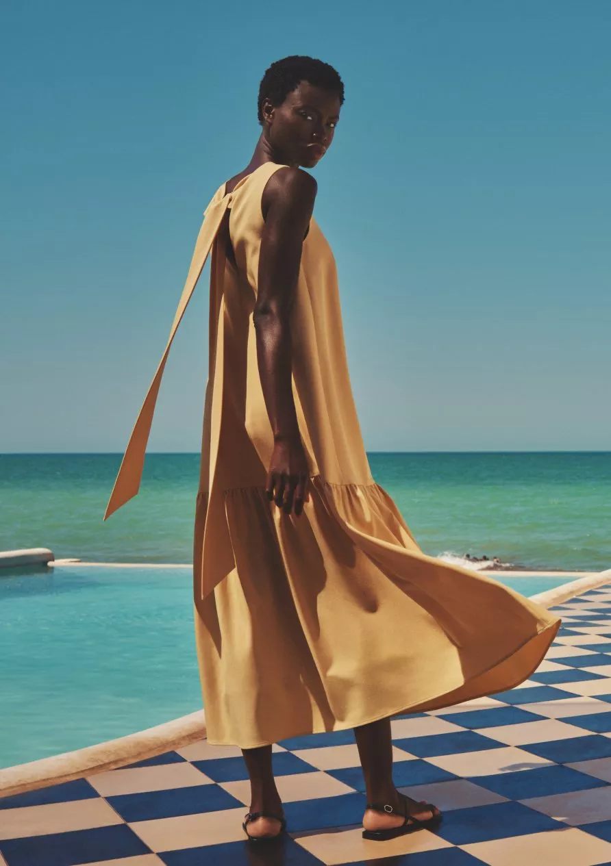 Fashion model in flowing yellow maxi dress by poolside with sea view, blue sky and tiled terrace backdrop